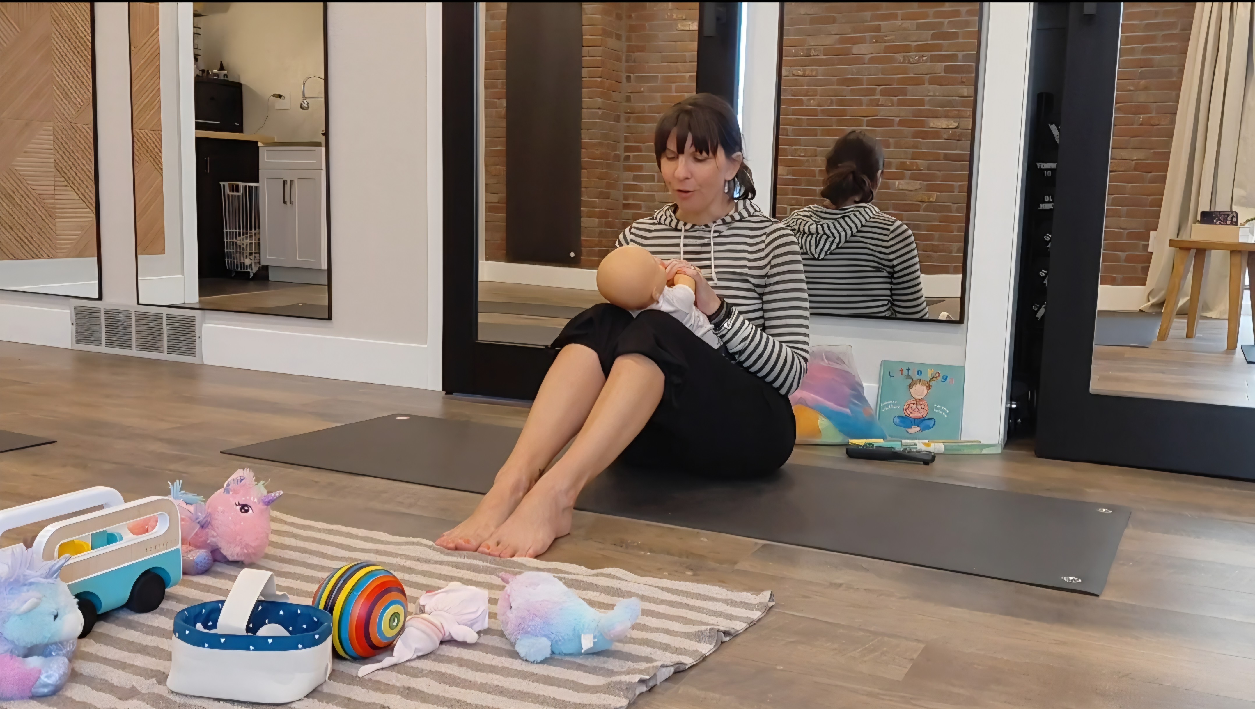 Woman sitting on a yoga mat in a room with toys and a striped shirt, holding a baby, with a mirror behind her.