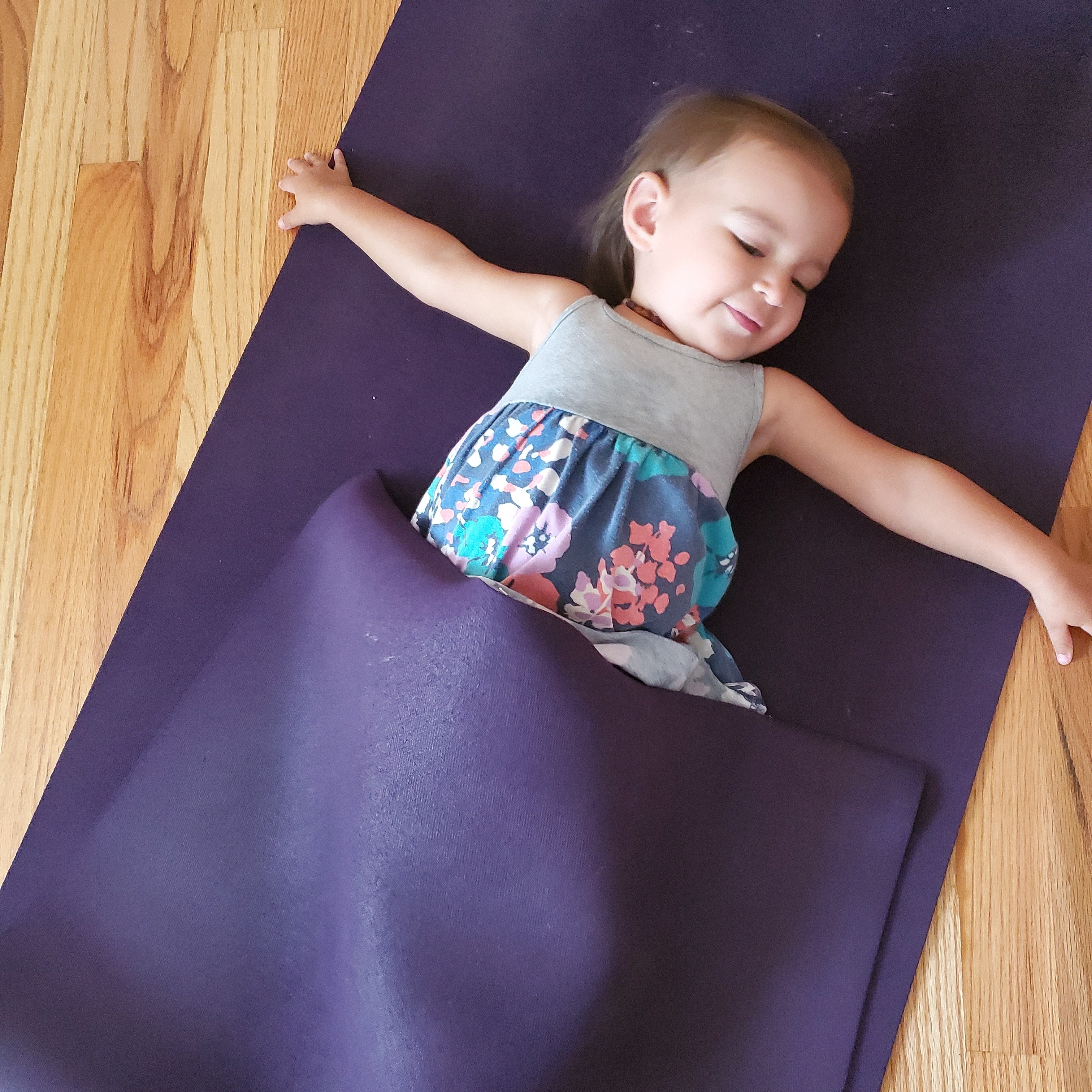 Child lying on a yoga mat with arms outstretched, partially covered by a blanket, on a wooden floor.