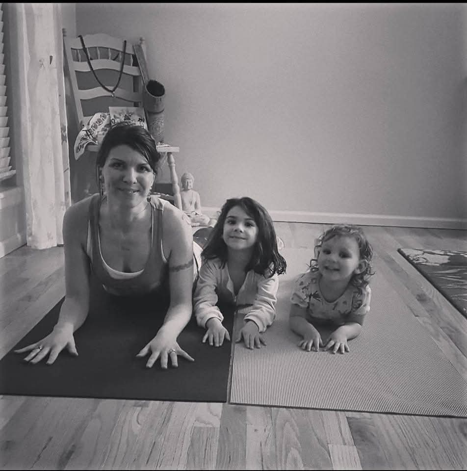 Woman and two children on yoga mats indoors, smiling, in a room with a wooden floor and furniture in the background.