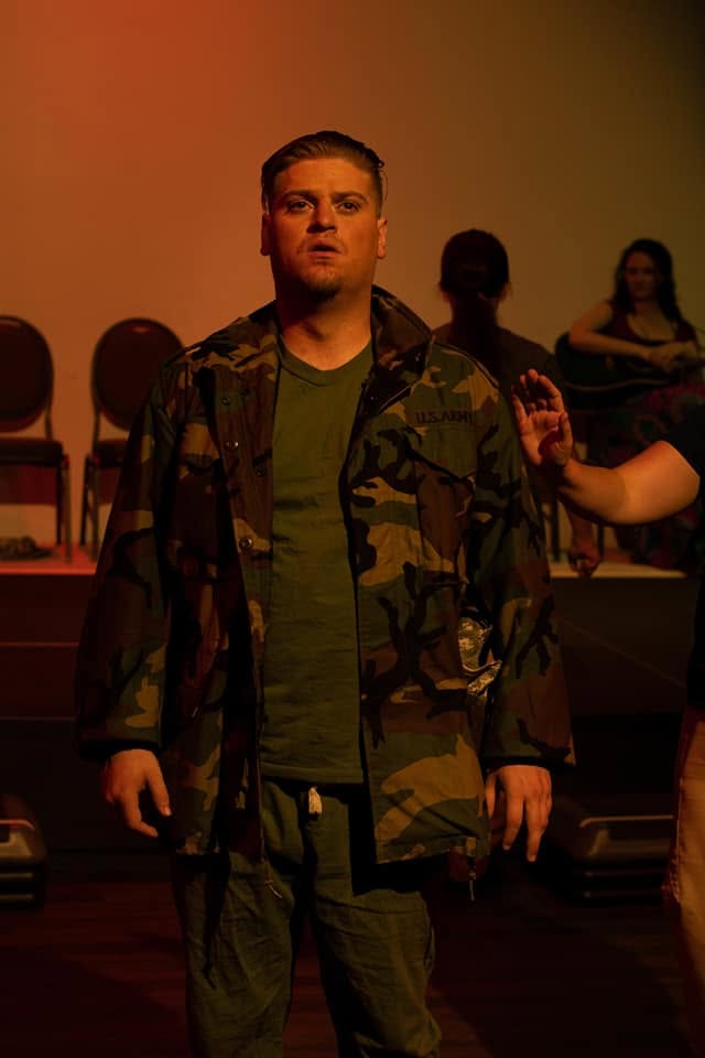 Man in camouflage jacket standing on stage with chairs and people in background.