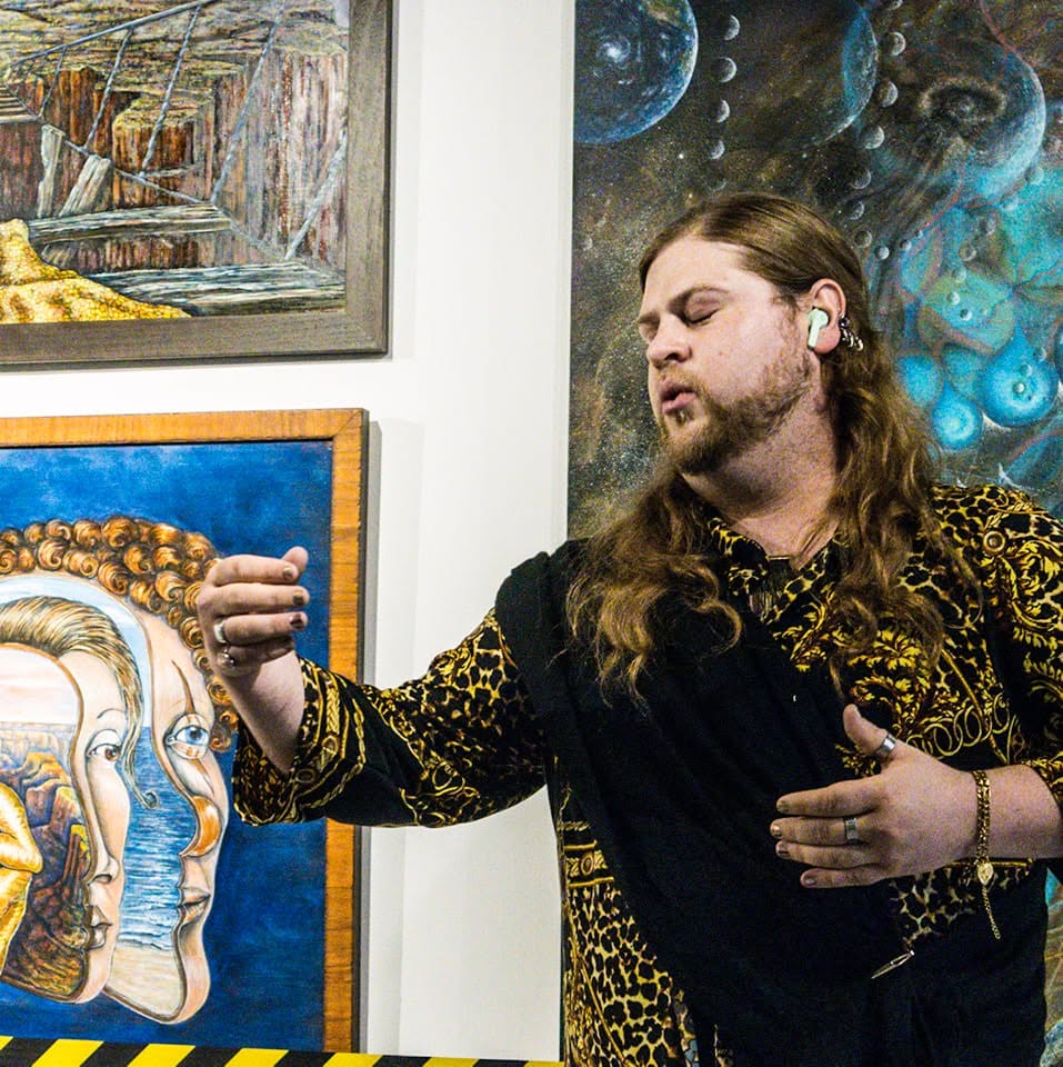 Man with long hair and a beard, wearing a leopard print jacket, stands in front of colorful artwork, gesturing with his hand.