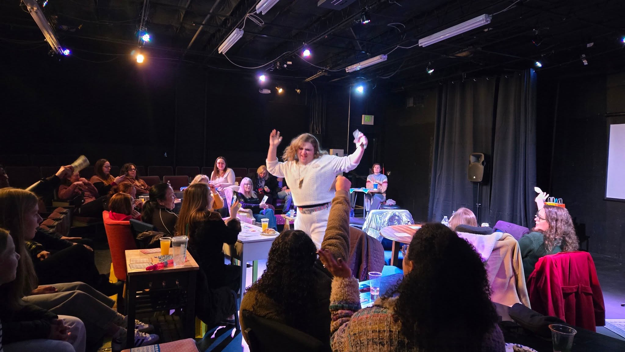 Person standing and speaking to an audience in a dark room with tables and chairs, some audience members raising hands.