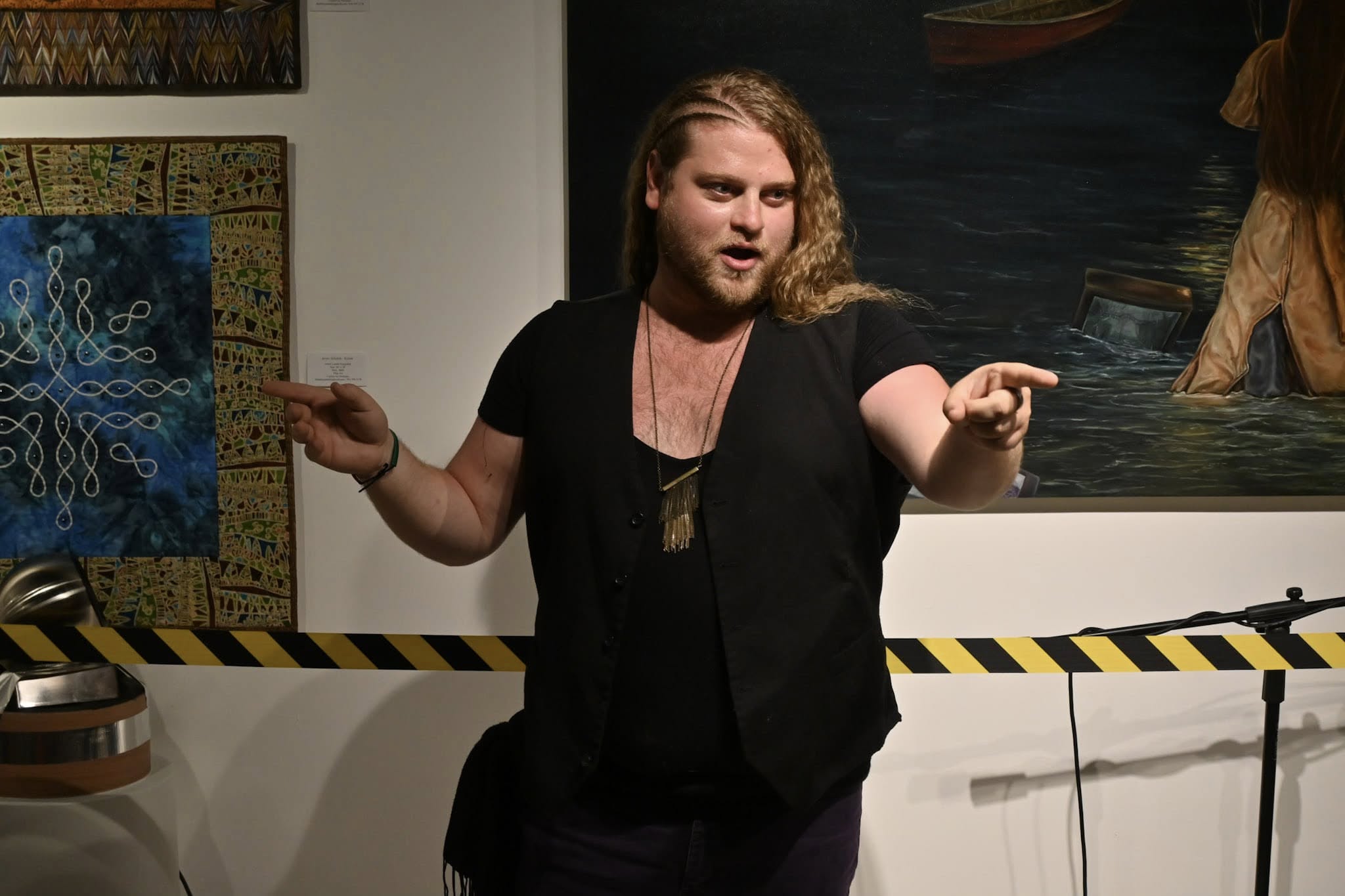 Person with long hair and beard gesturing with hands in an art gallery, paintings on the wall behind.