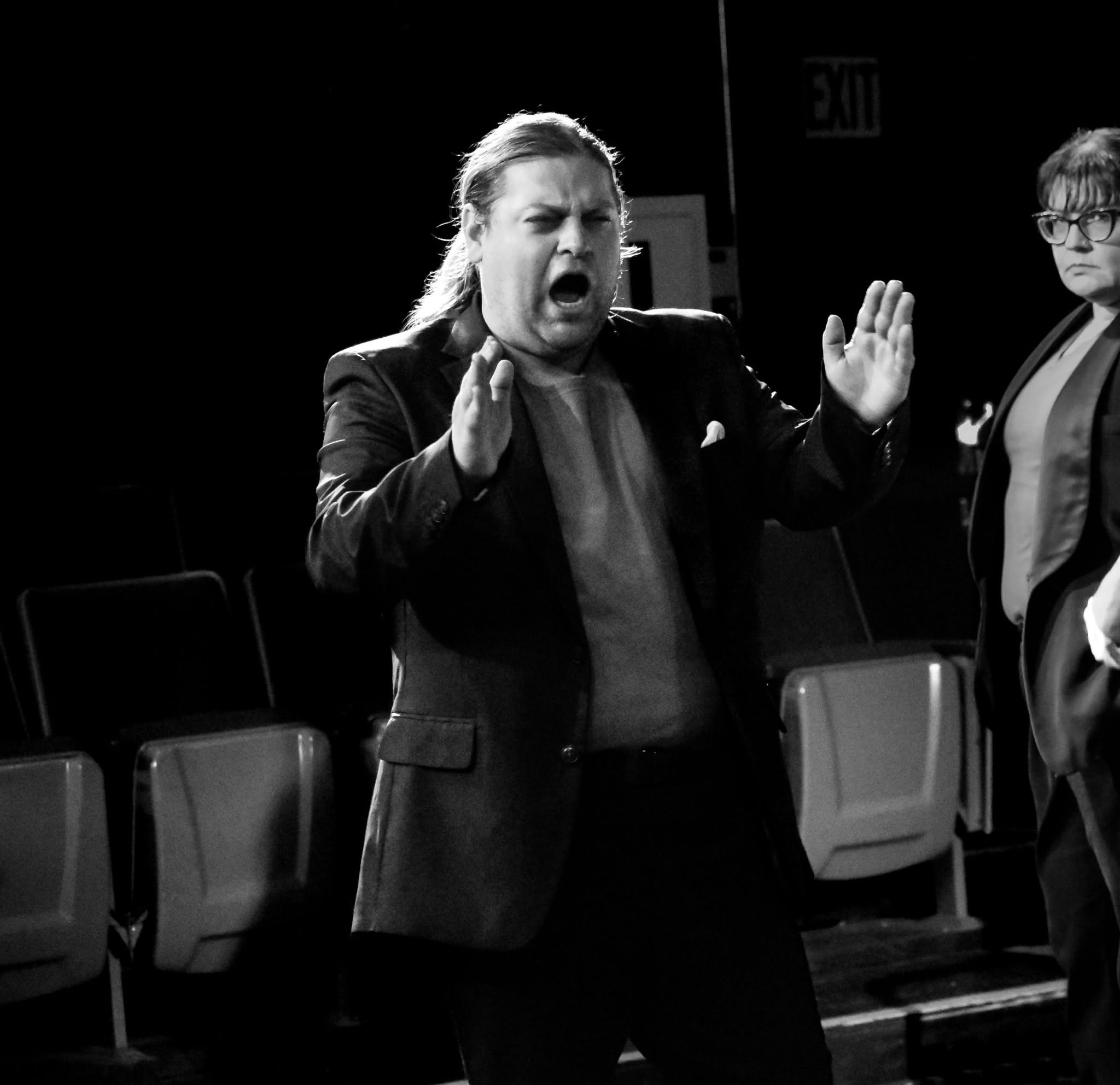 Person with long hair and open mouth gesturing with hands, in a dark room with chairs, black and white photo.