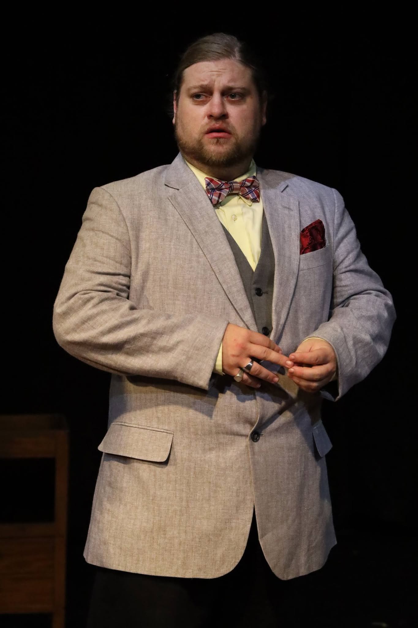 Man in a light gray suit with a bow tie, holding a small object, standing against a black background.