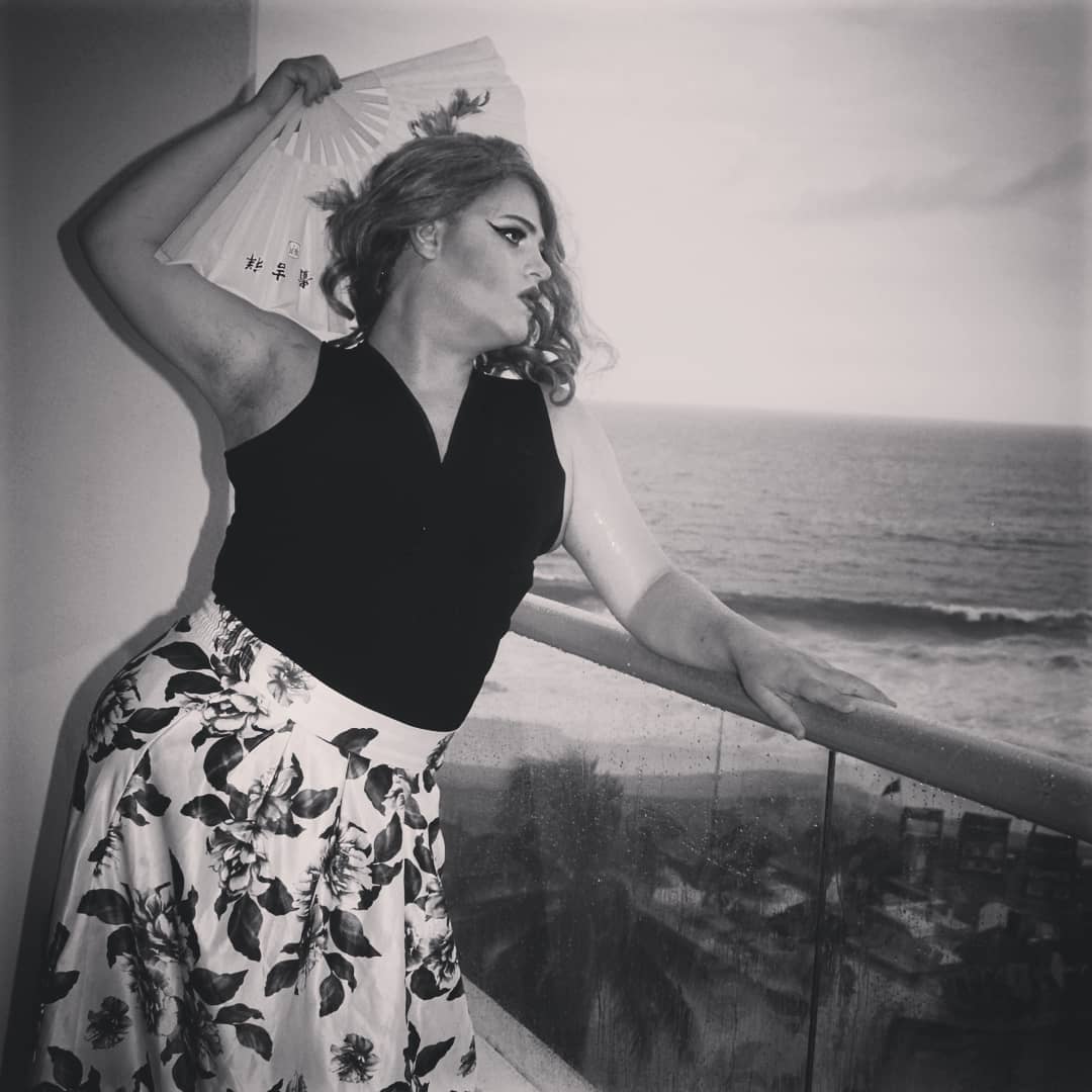 Woman holding a fan, looking to the side, standing on a balcony overlooking the beach, in black and white.