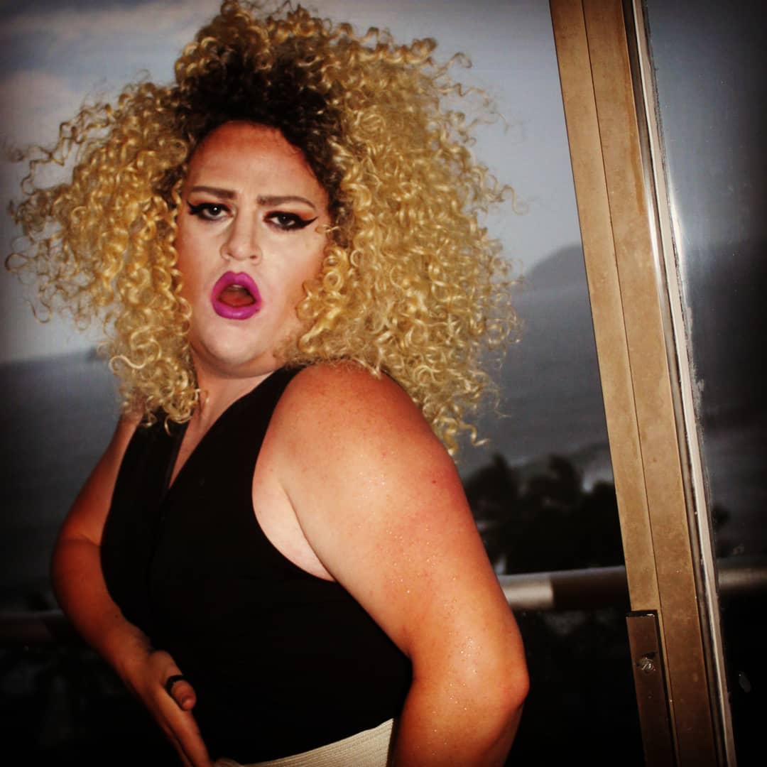 Person with curly blonde hair and bold makeup, wearing a black top, standing outdoors near a glass door.