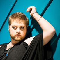 Young man with short hair and beard, wearing a black shirt, looking serious, against a blue background with diagonal lines.