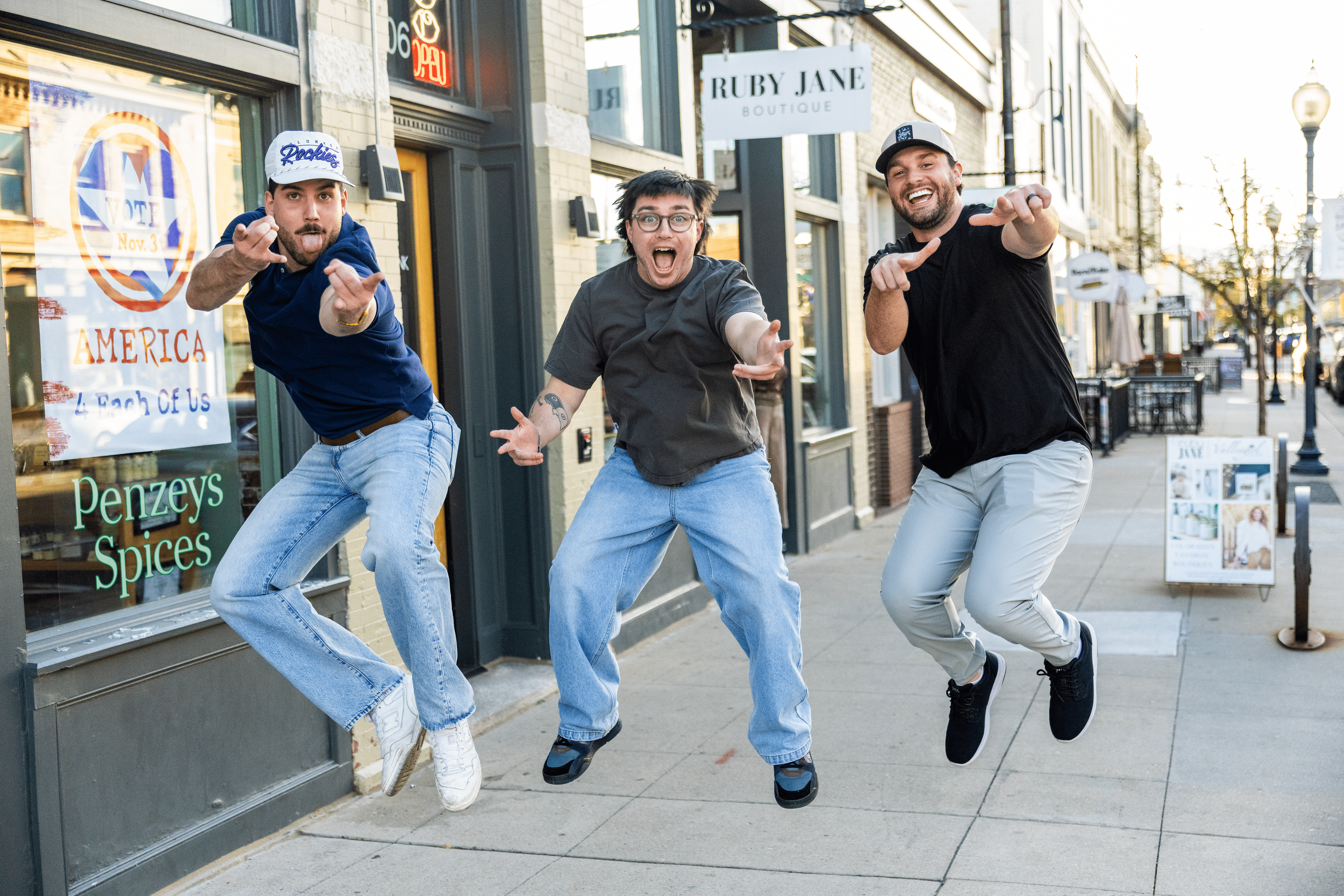Three people jumping and pointing at the camera outside a storefront on a city sidewalk.