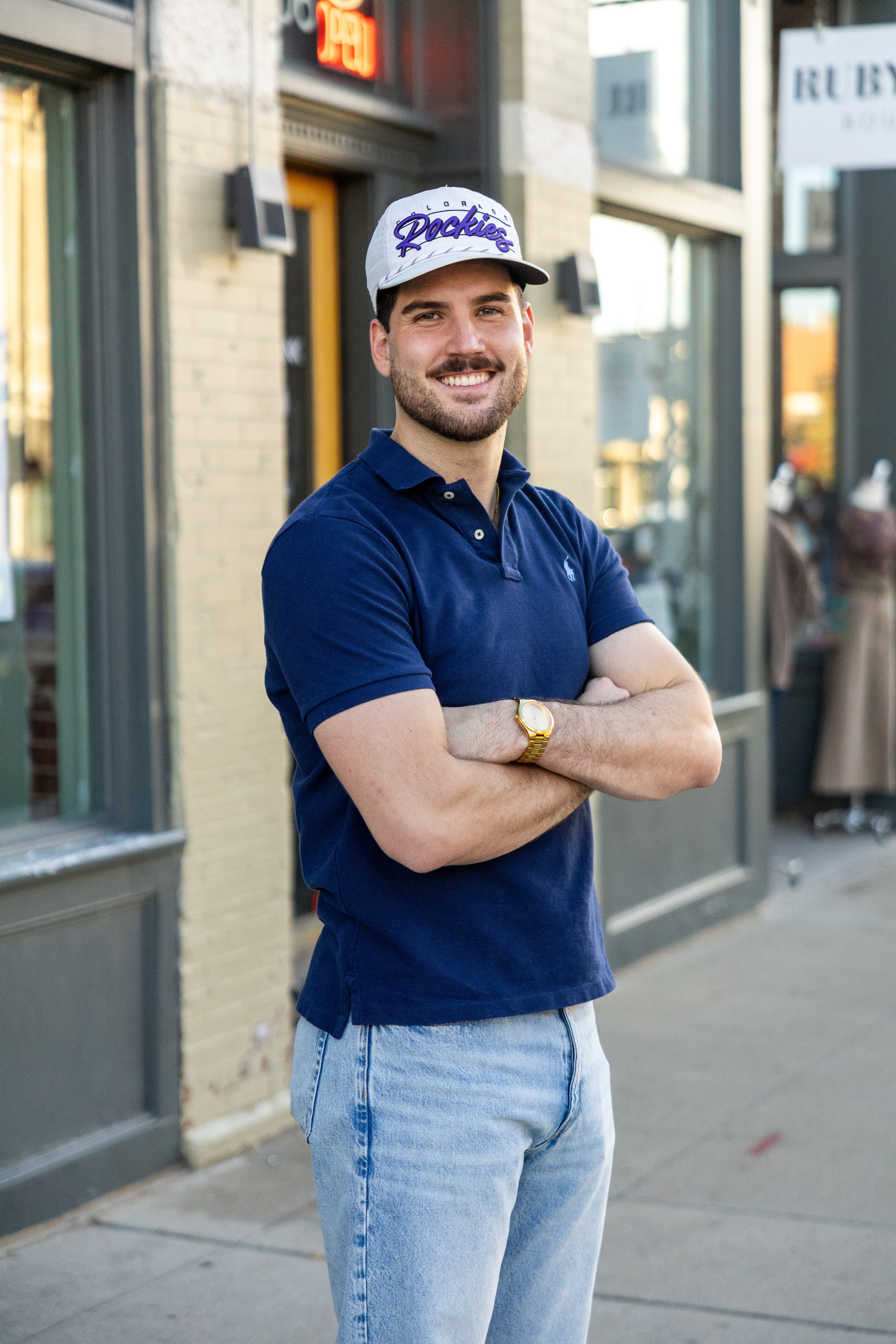 Smiling man with crossed arms wearing a white cap, navy polo shirt, and light jeans, standing outside a building.