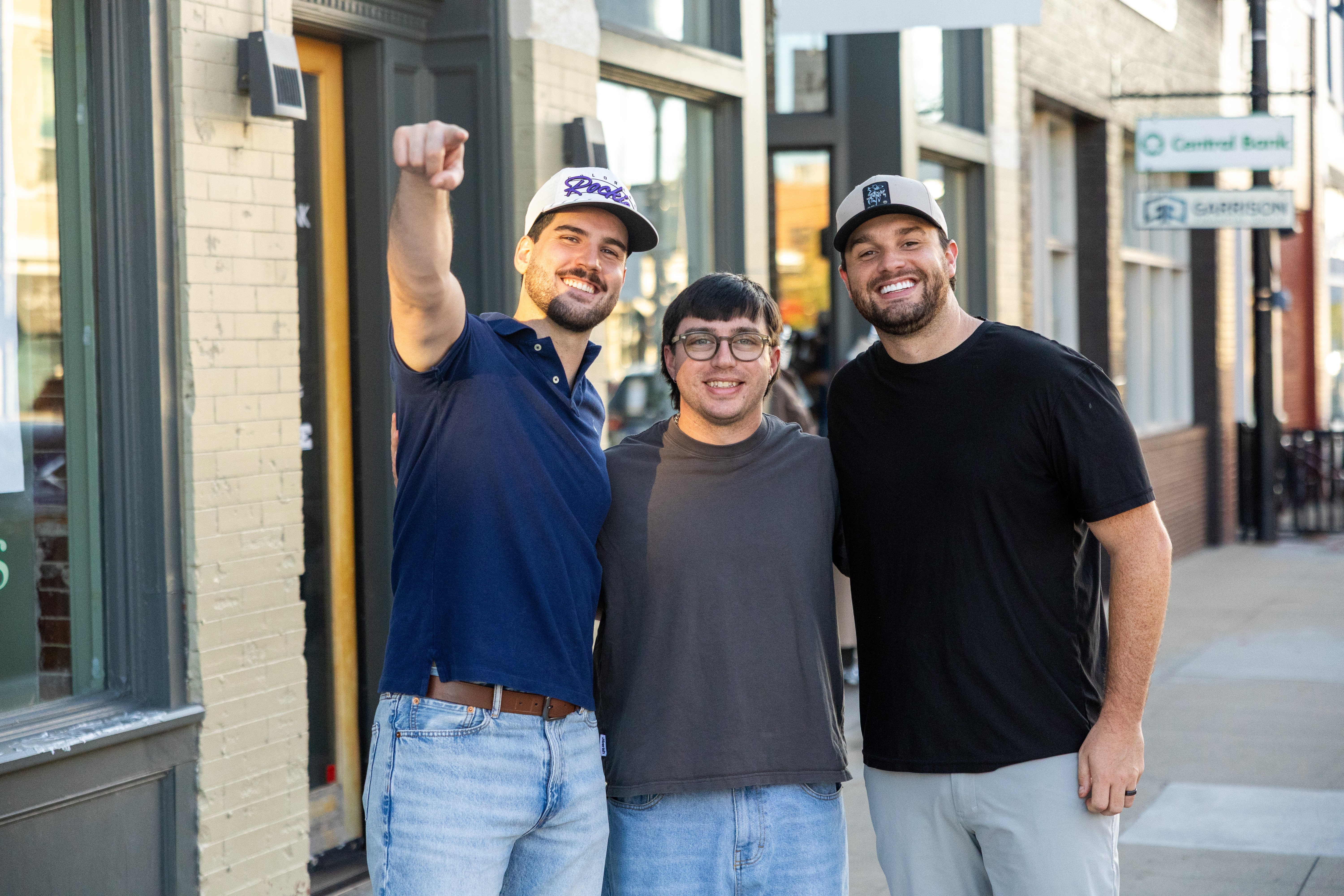 Three smiling people standing outside on a sidewalk, two men and a young person, with buildings in the background.