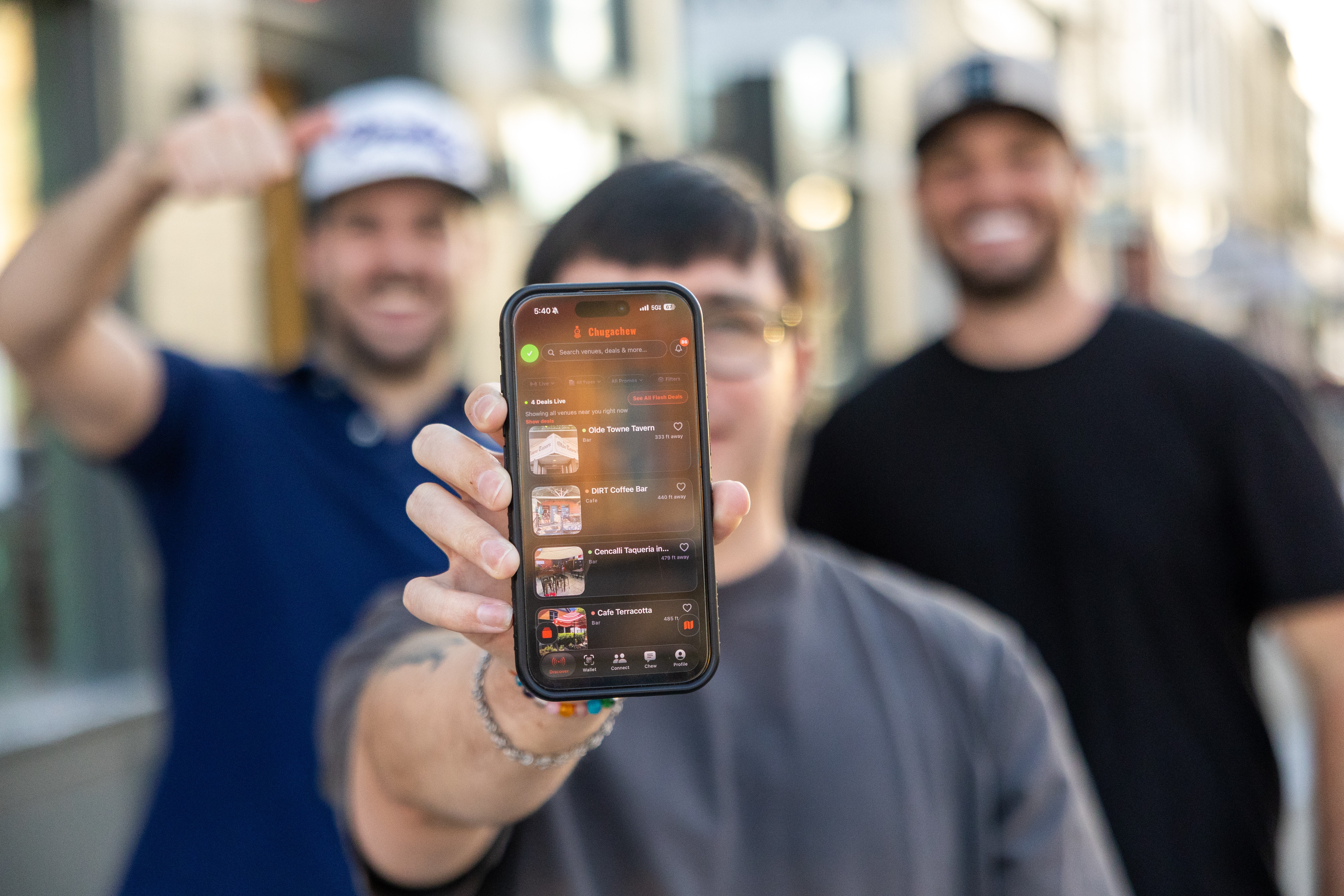 Person holding smartphone in front of two smiling people outdoors, blurred background, sunny day.