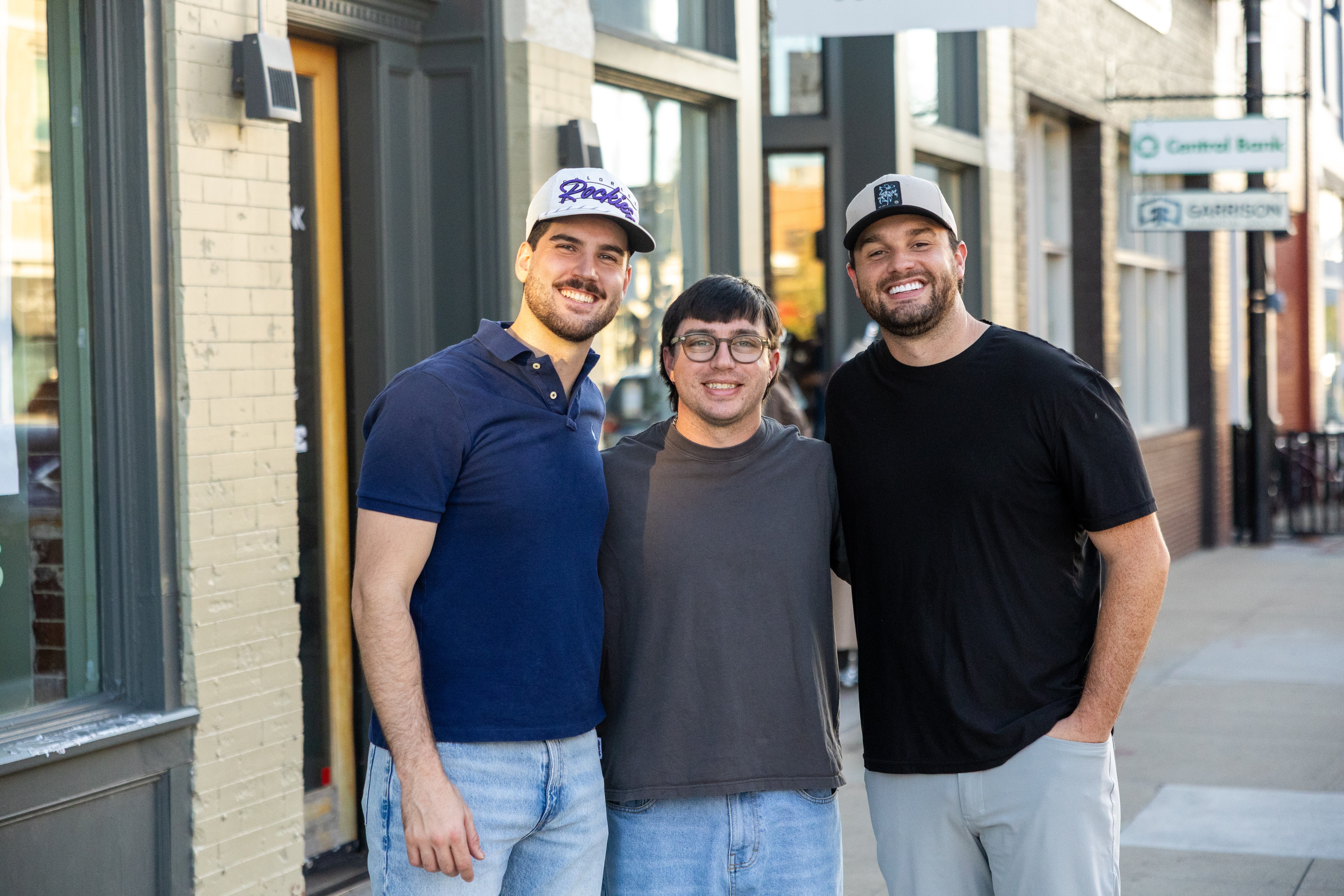 Three people standing outside a building, smiling, with storefronts in the background.