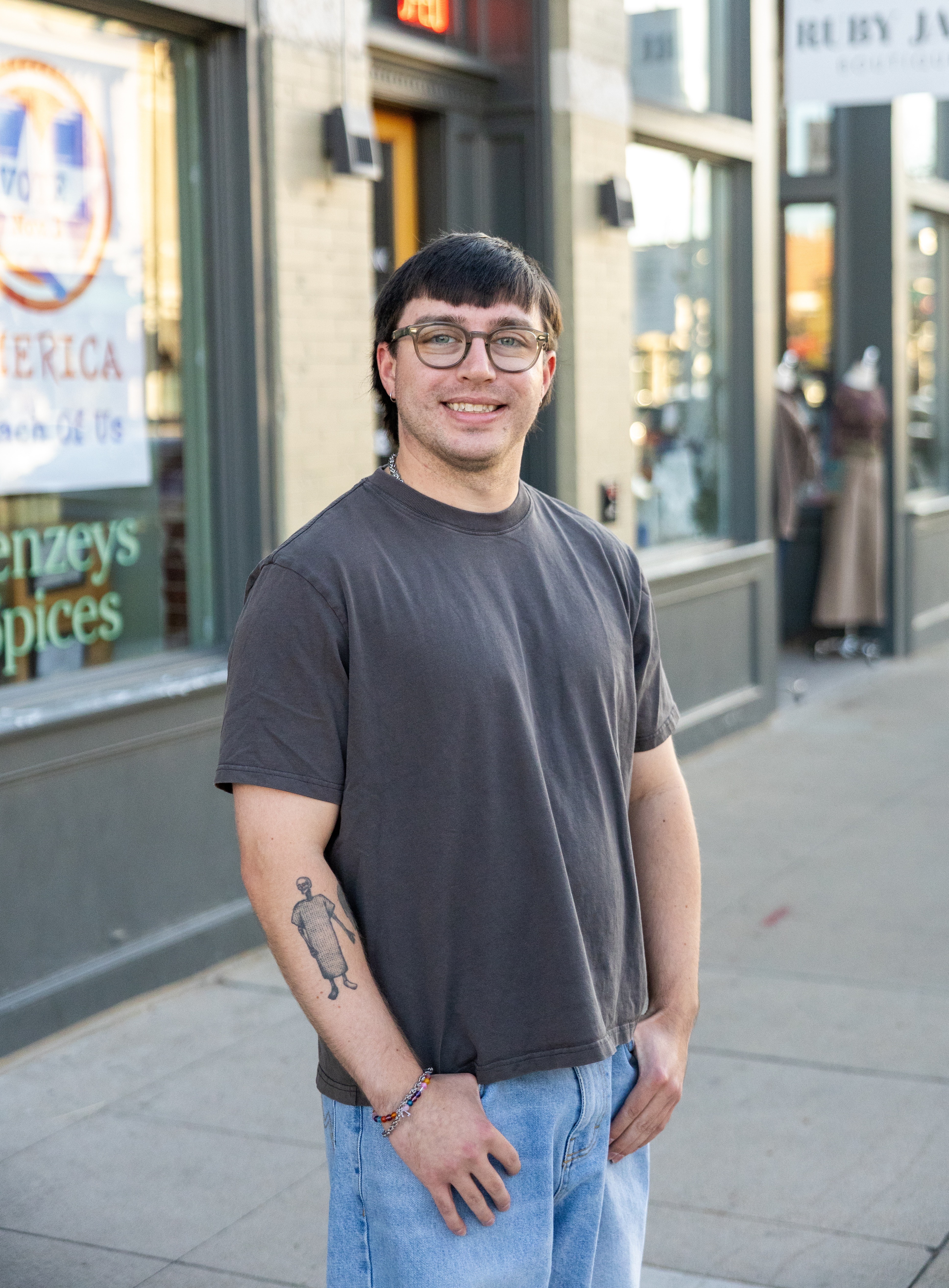 Young man with glasses and a tattoo on his left arm stands on a city sidewalk smiling, wearing a dark t-shirt and jeans.