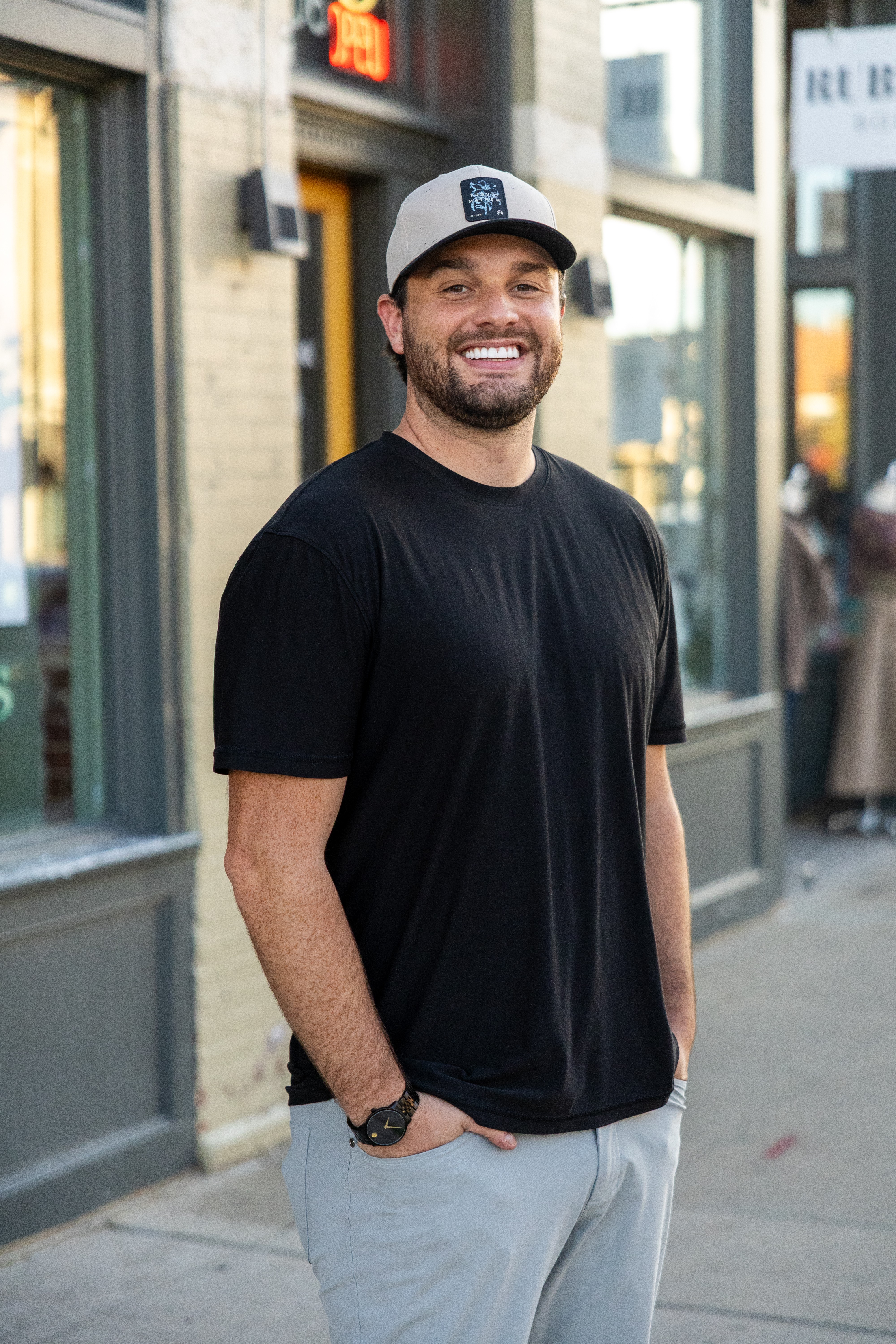 Smiling man with a beard wearing a black t-shirt, white cap, and light-colored pants standing on a city sidewalk.