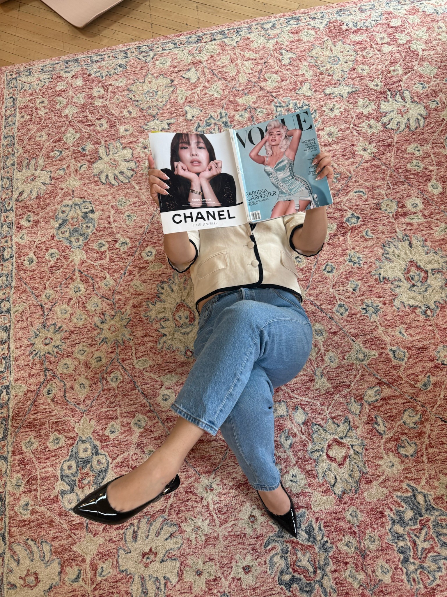 Person lying on a pink patterned rug, holding two magazine pages with images of women, wearing beige top and blue jeans.