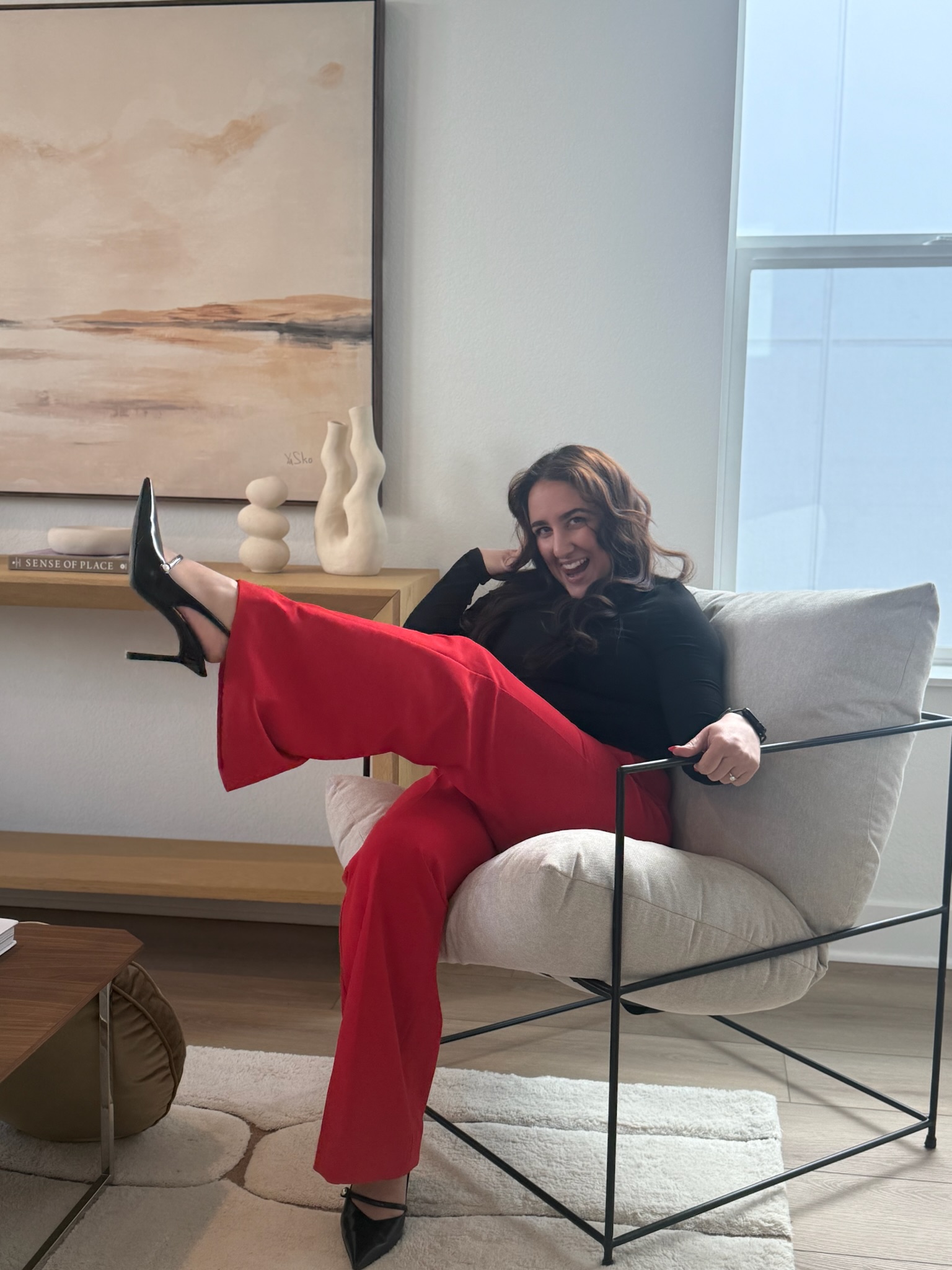 Woman sitting on a modern chair with a high back, smiling, wearing red pants and black top, in a bright room.