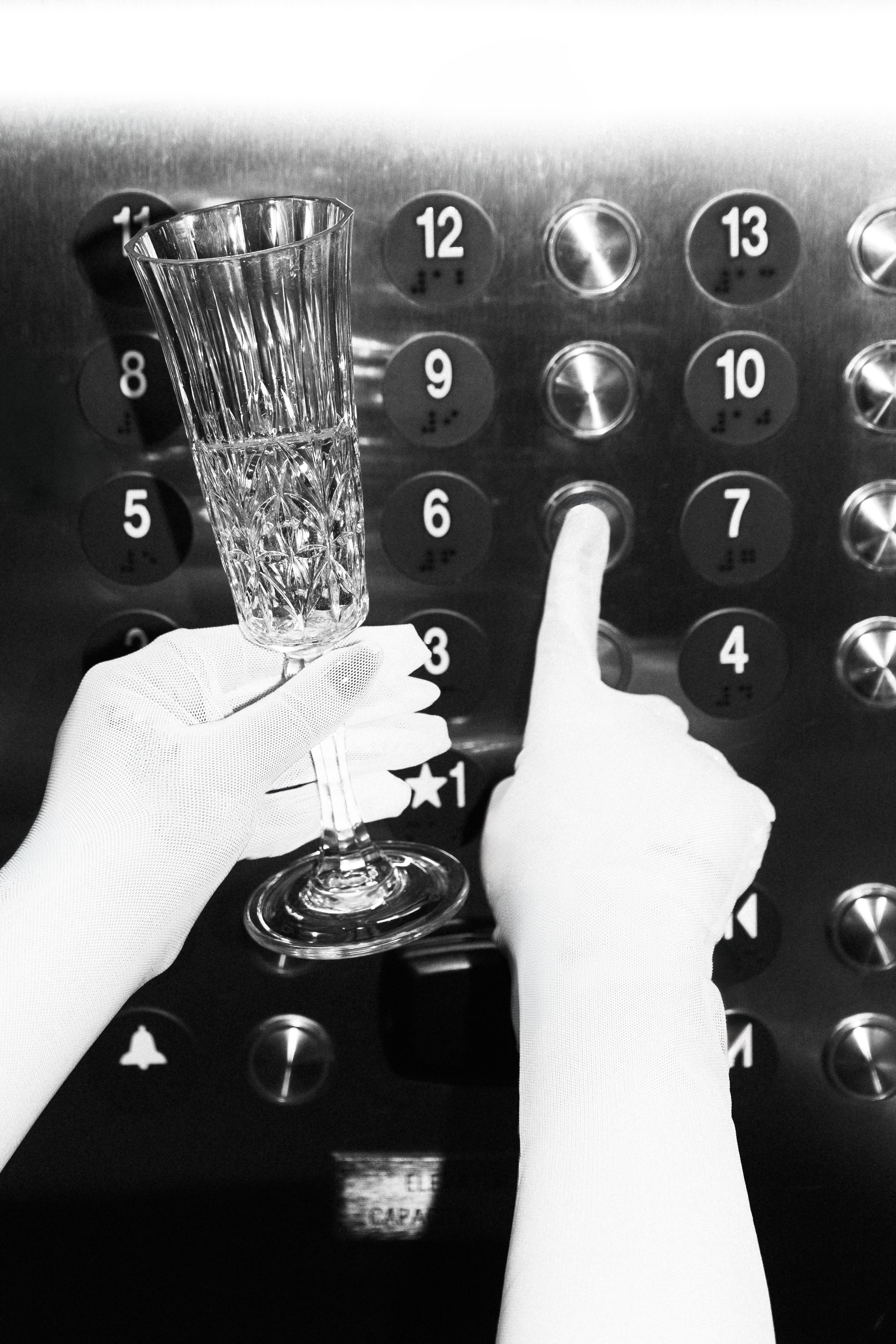Person's hand holding a glass of champagne near elevator buttons, with one finger pointing at button 3.
