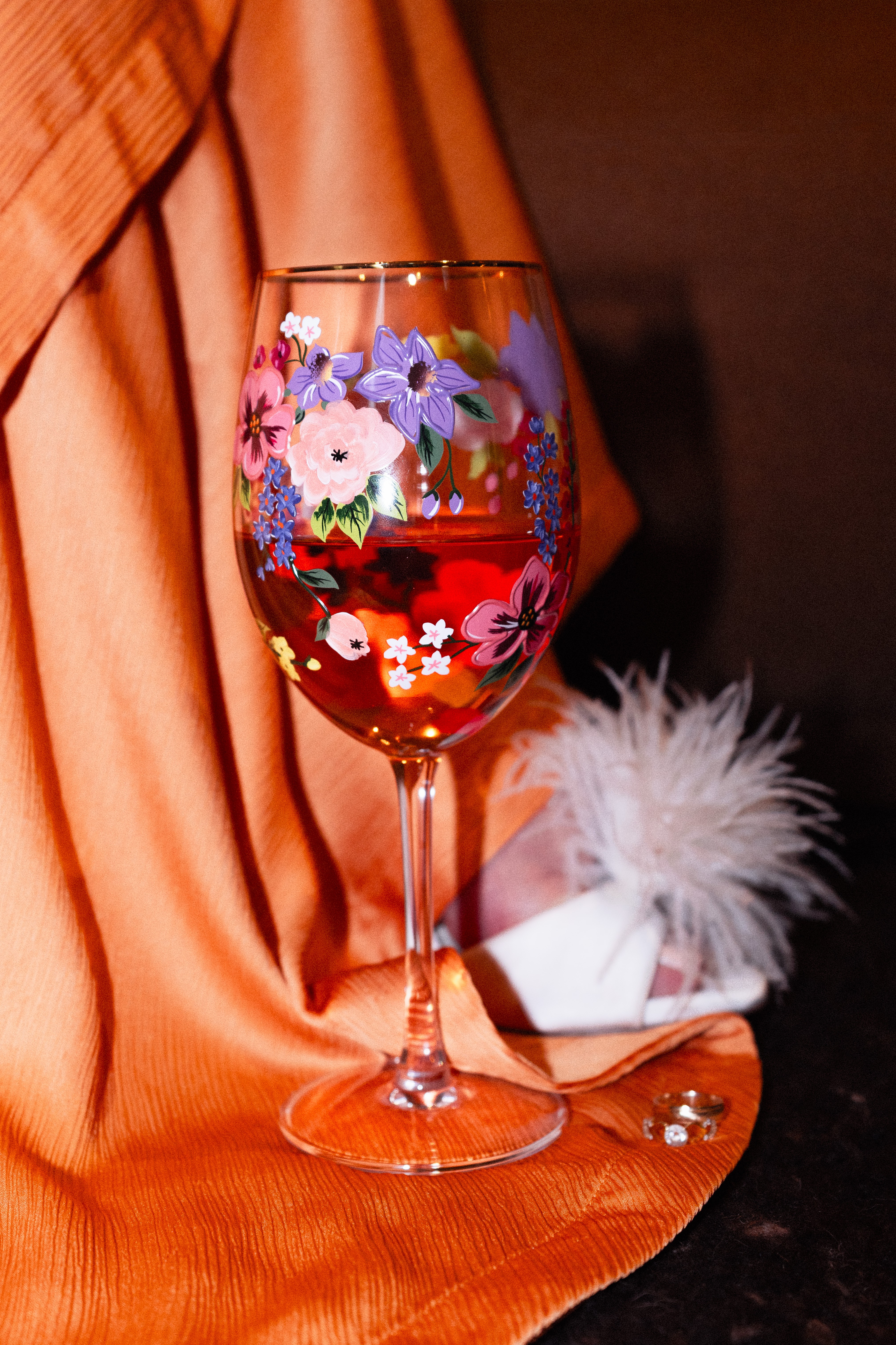 A glass of red wine with floral designs, placed on an orange fabric surface with a white feathered accessory nearby.