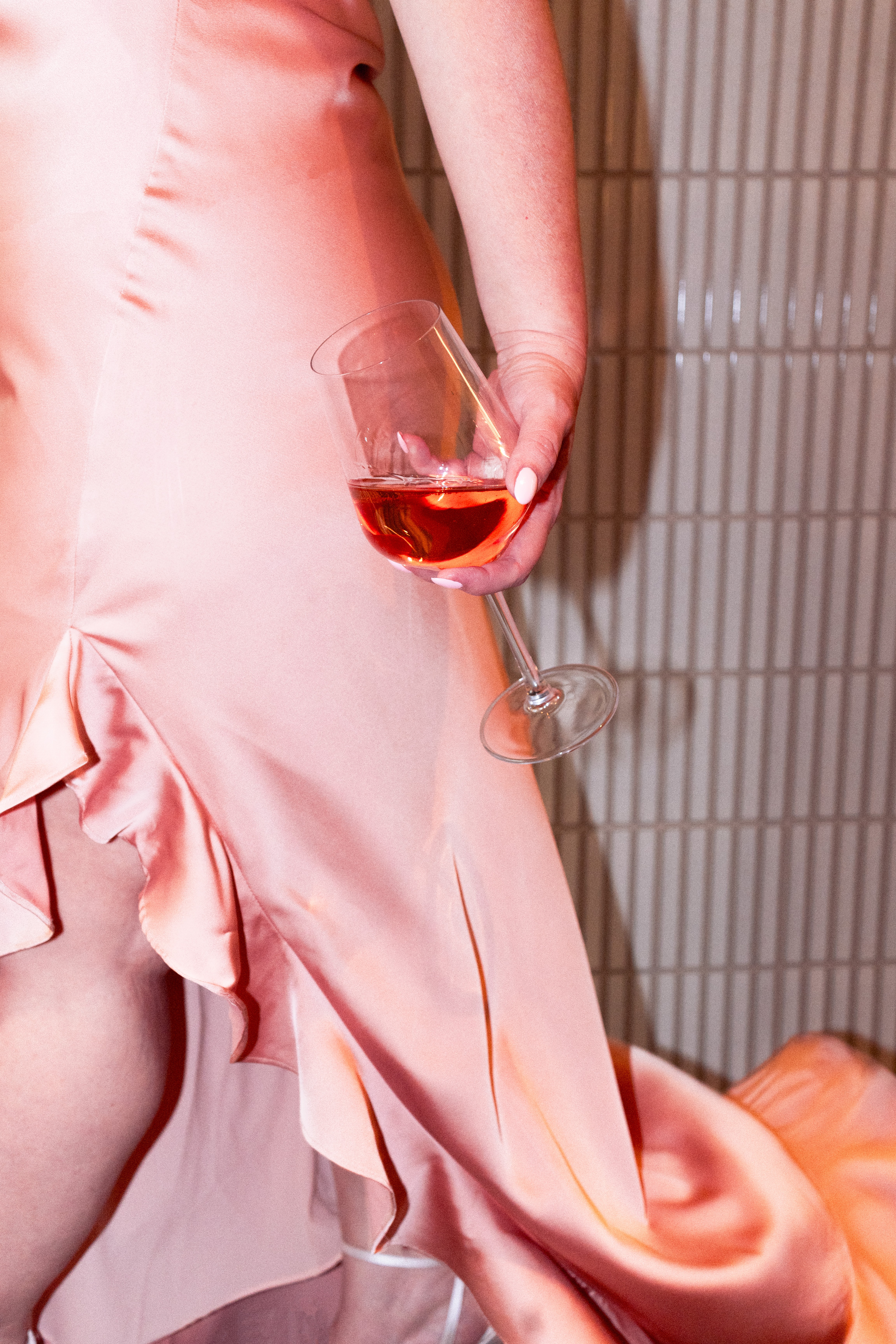 Person in pink dress holding a glass of pink wine, standing near a tiled wall.