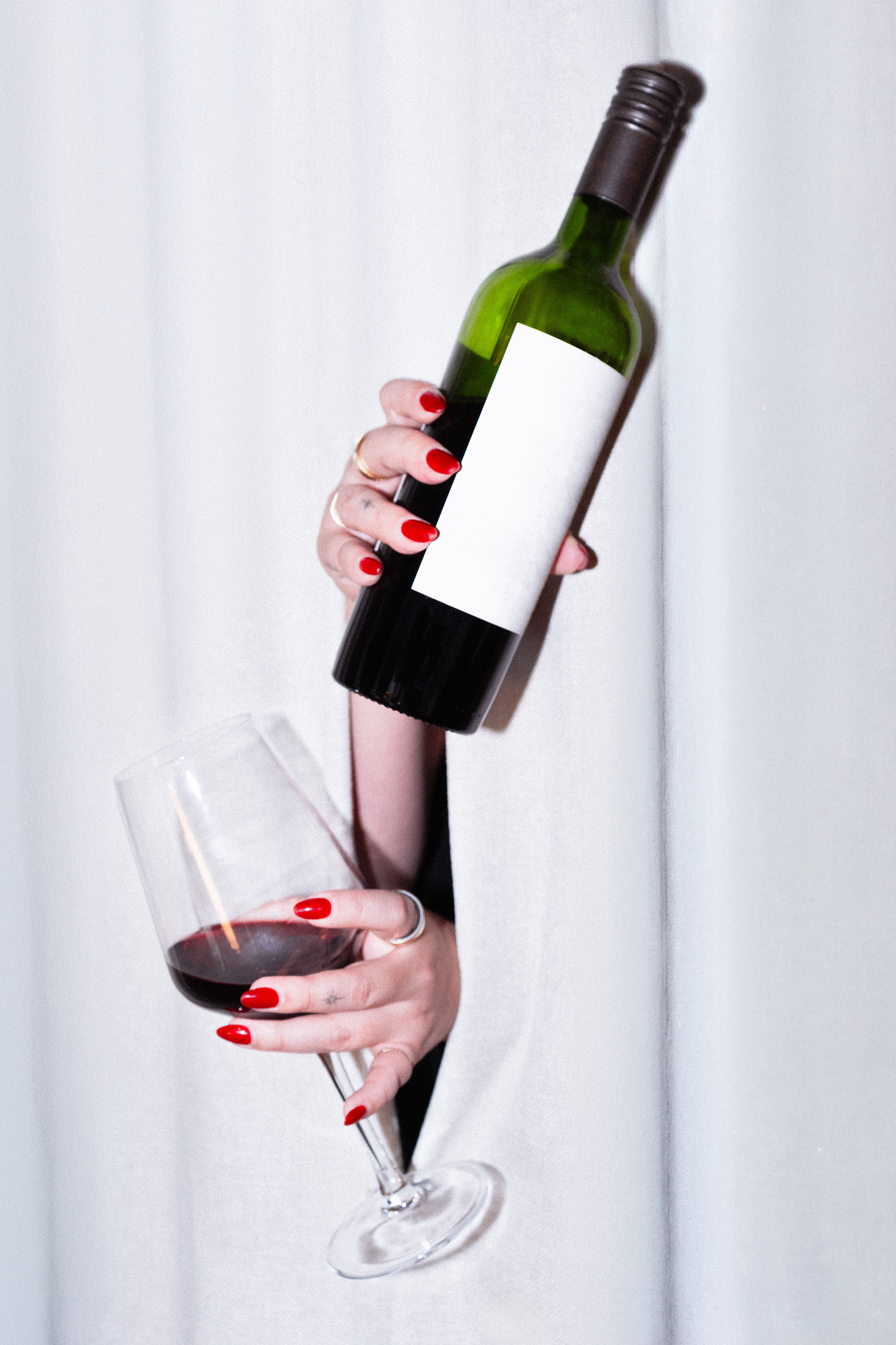 Person holding a wine bottle and glass with red wine, against a white background, with red nail polish.