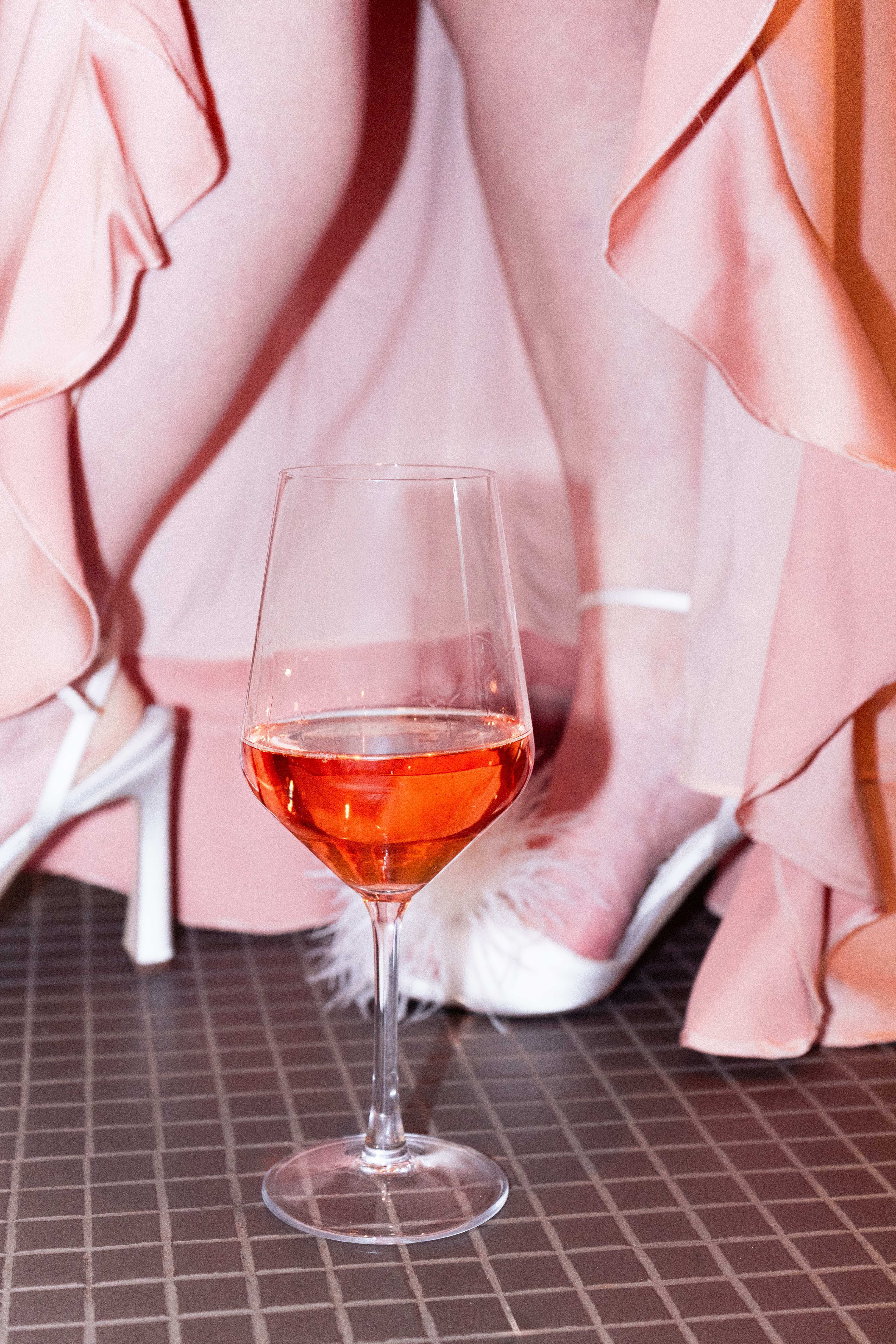 Person in pink clothing with ruffled sleeves, holding a glass of pink wine, with a white shoe visible.