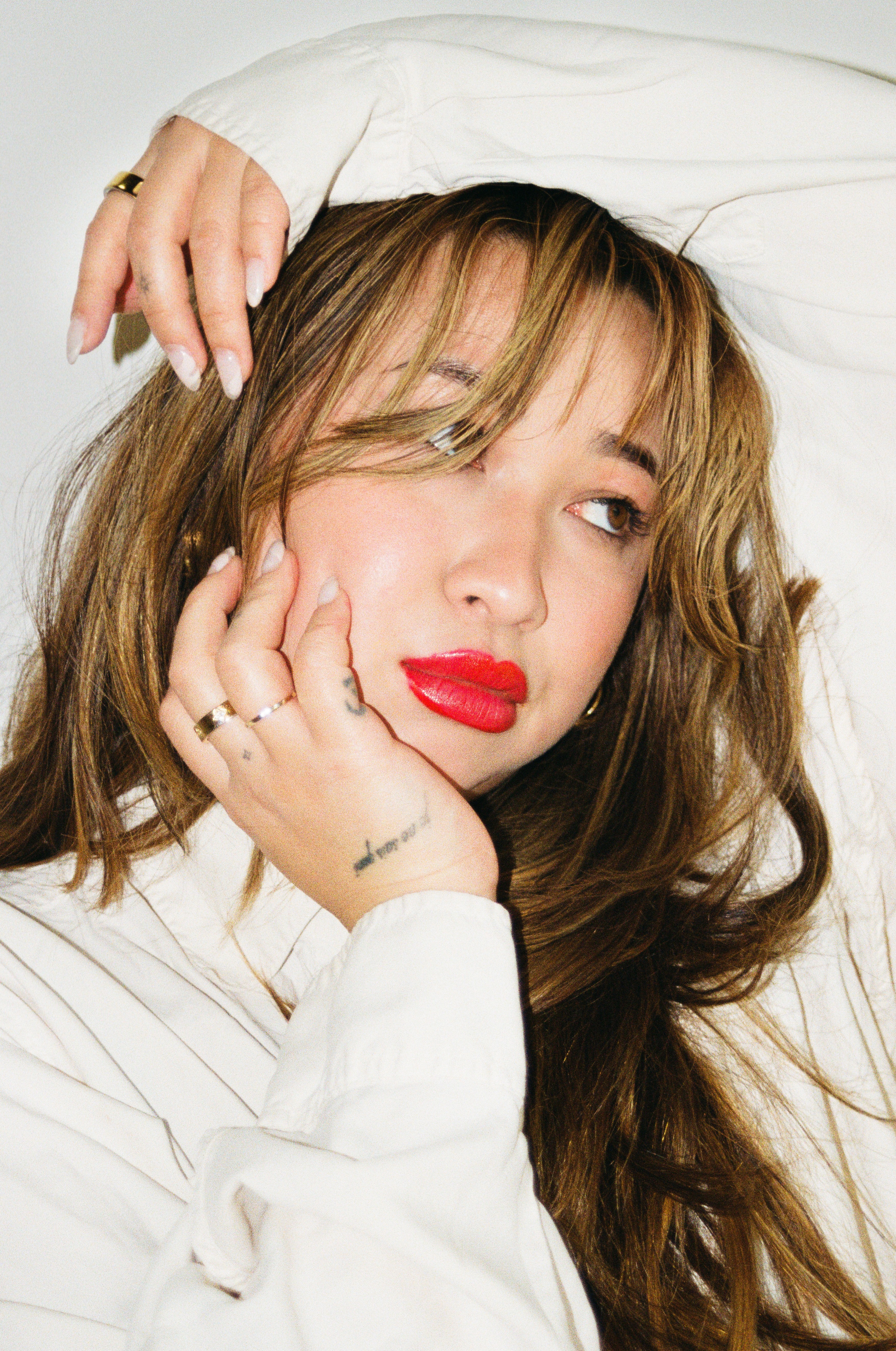 Woman with long hair, red lipstick, resting her face on her hand, wearing a white top, looking to the side.