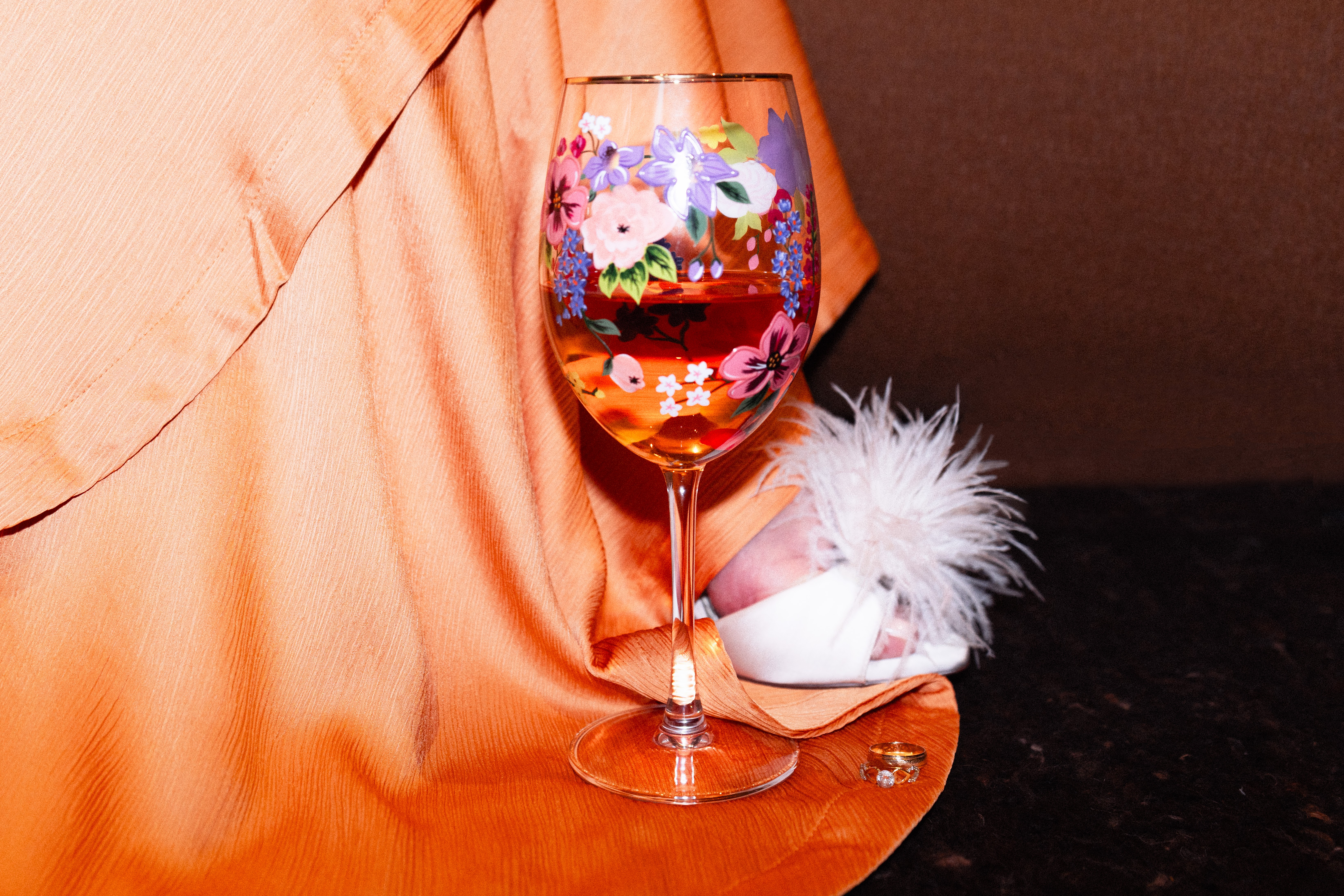 A glass of colorful flower-shaped candies on a table with a peach-colored cloth, a white feathered object, and a brown background.