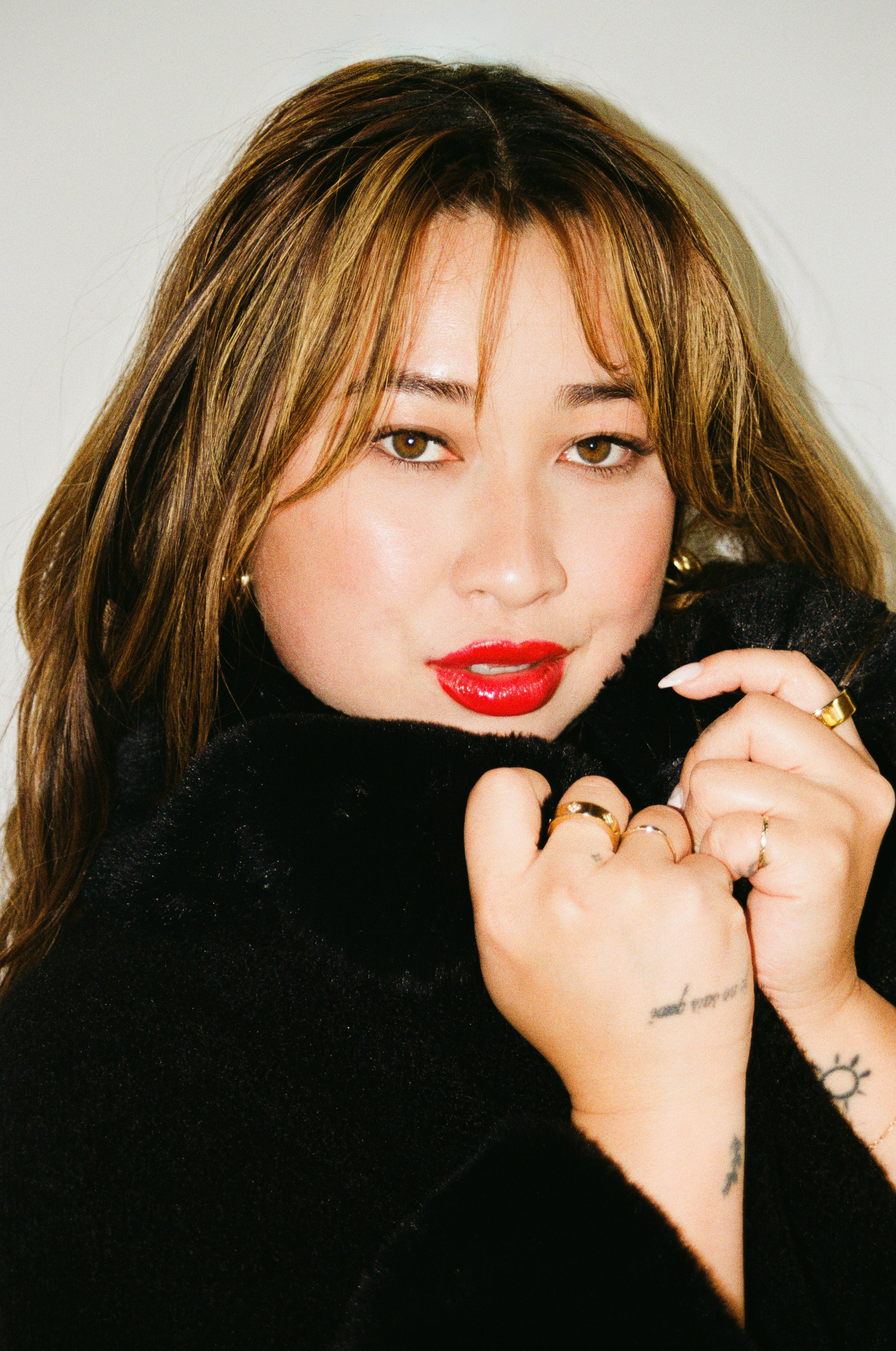 Woman with light brown hair, red lipstick, wearing a black outfit, holding her collar close, looking at the camera.