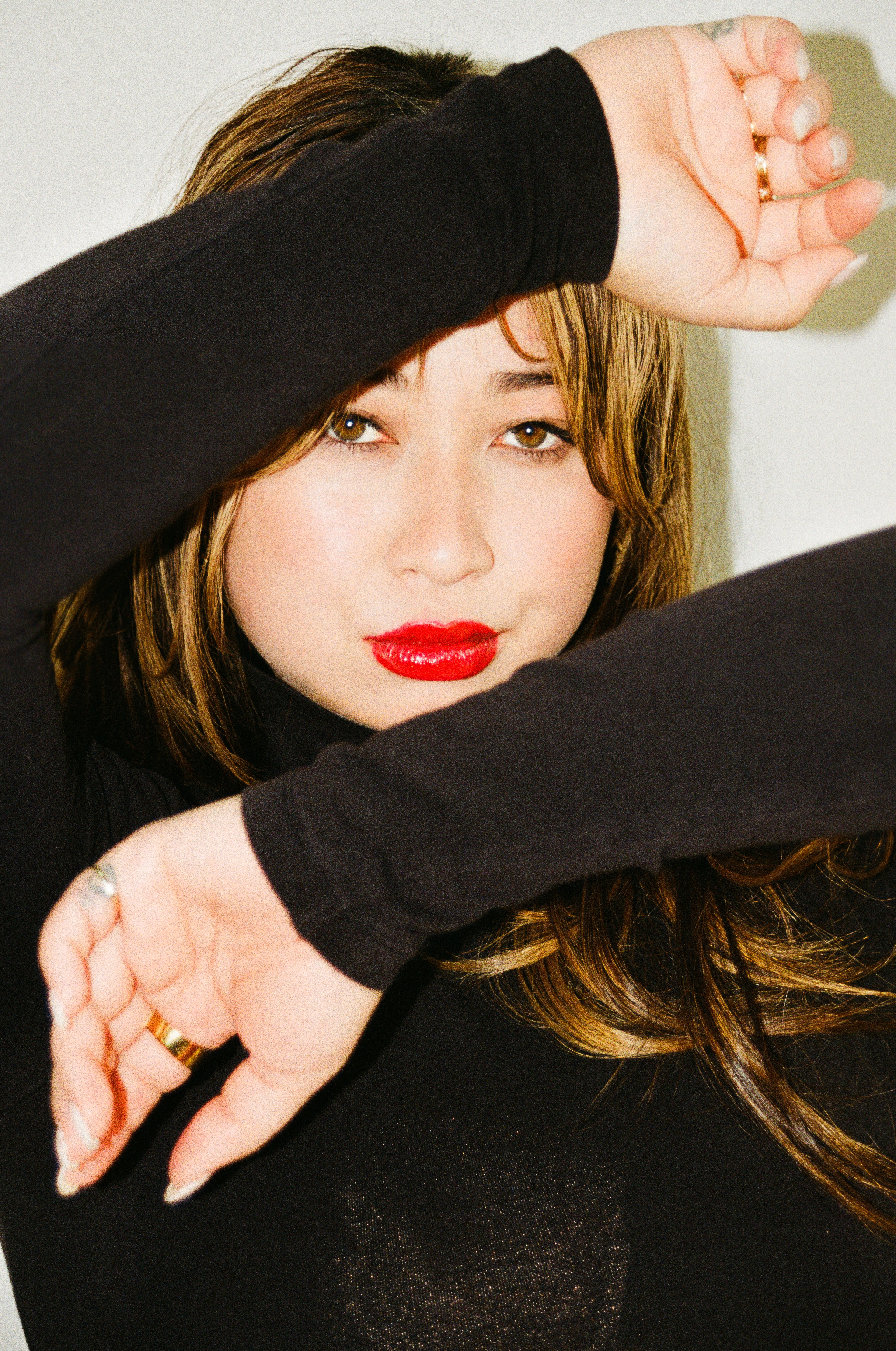 Young woman with light skin, red lipstick, and brown hair, wearing a black top, covering part of her face with her arm.