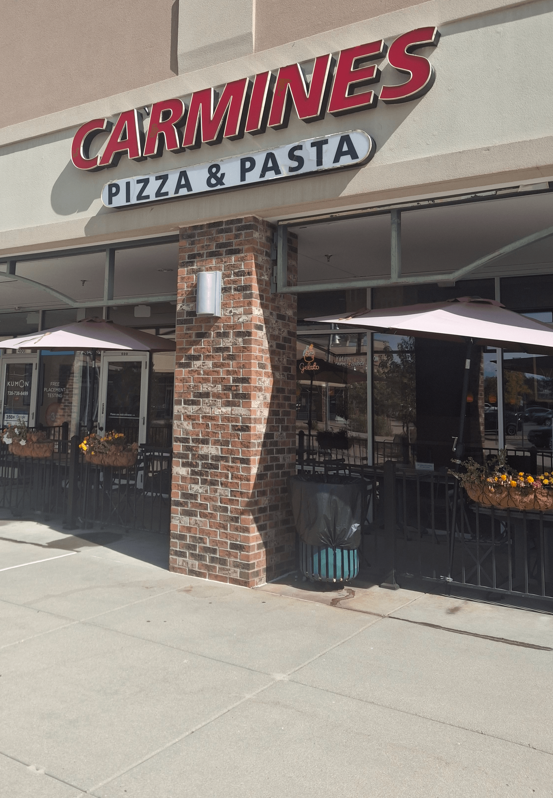 Carmine's Pizza & Pasta restaurant exterior with brick pillar, outdoor seating, and umbrellas.
