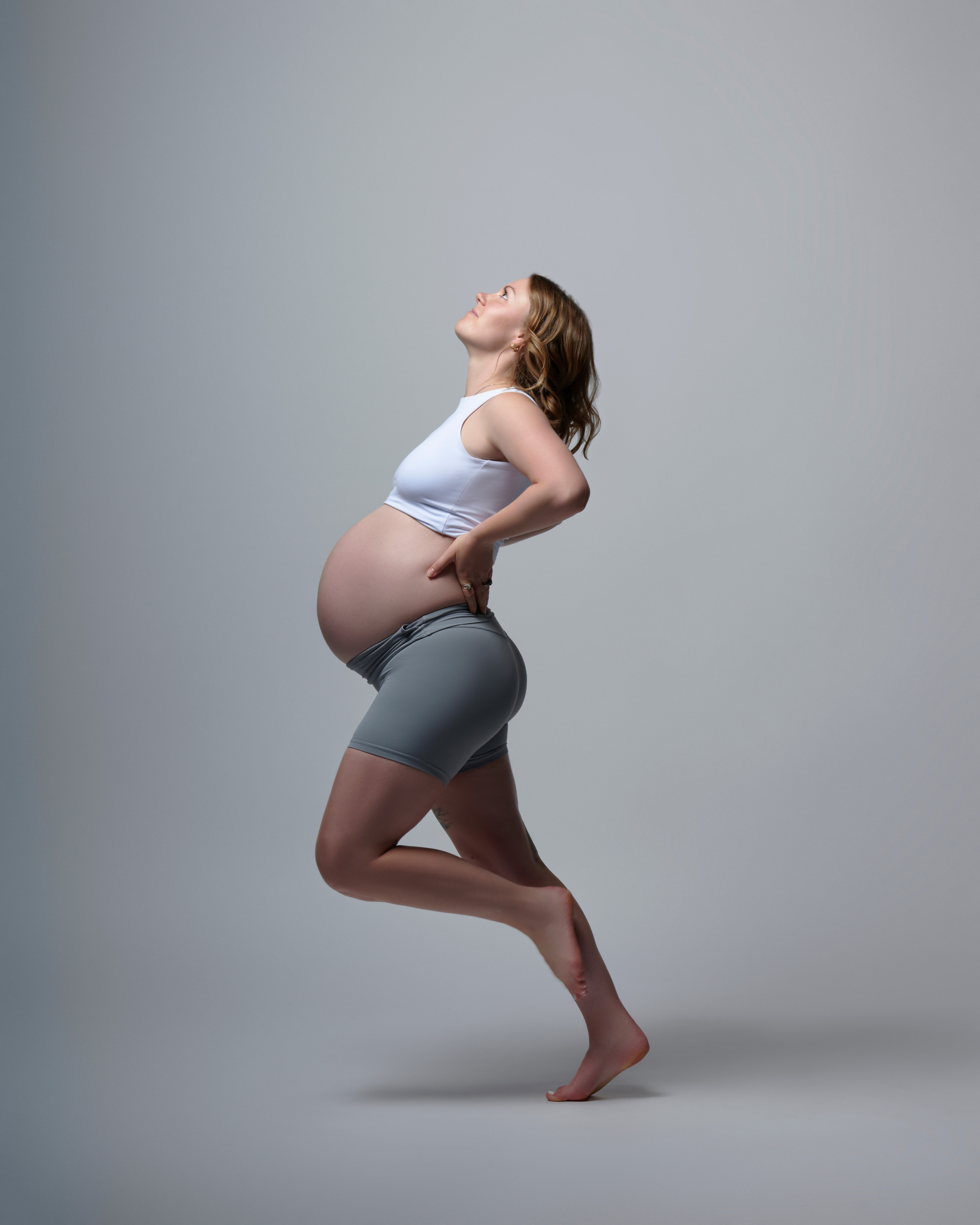 Pregnant woman in athletic wear standing on one leg with arms behind her back, head tilted back, smiling.