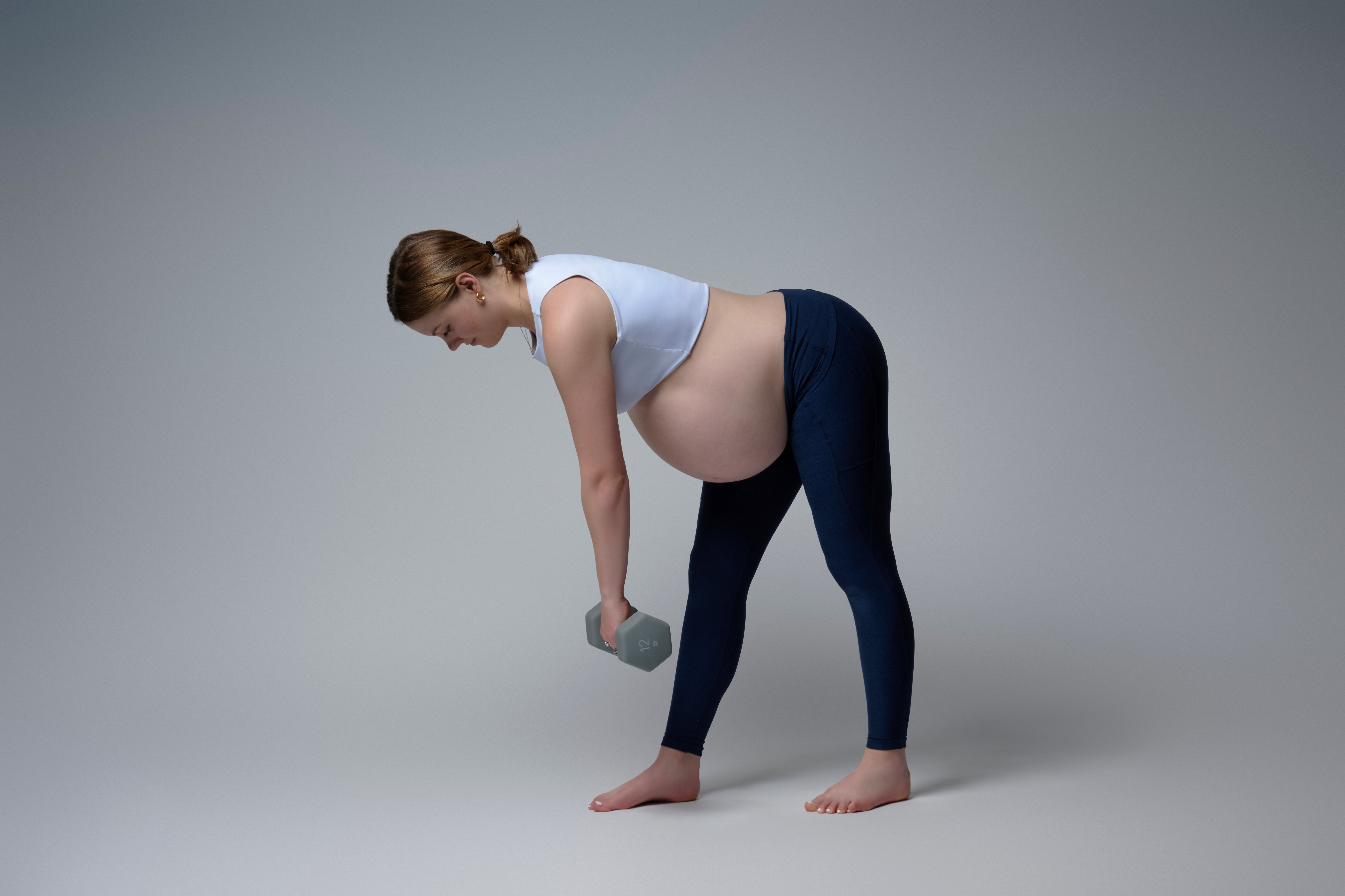Pregnant woman in workout clothes bending forward holding a dumbbell, gray background