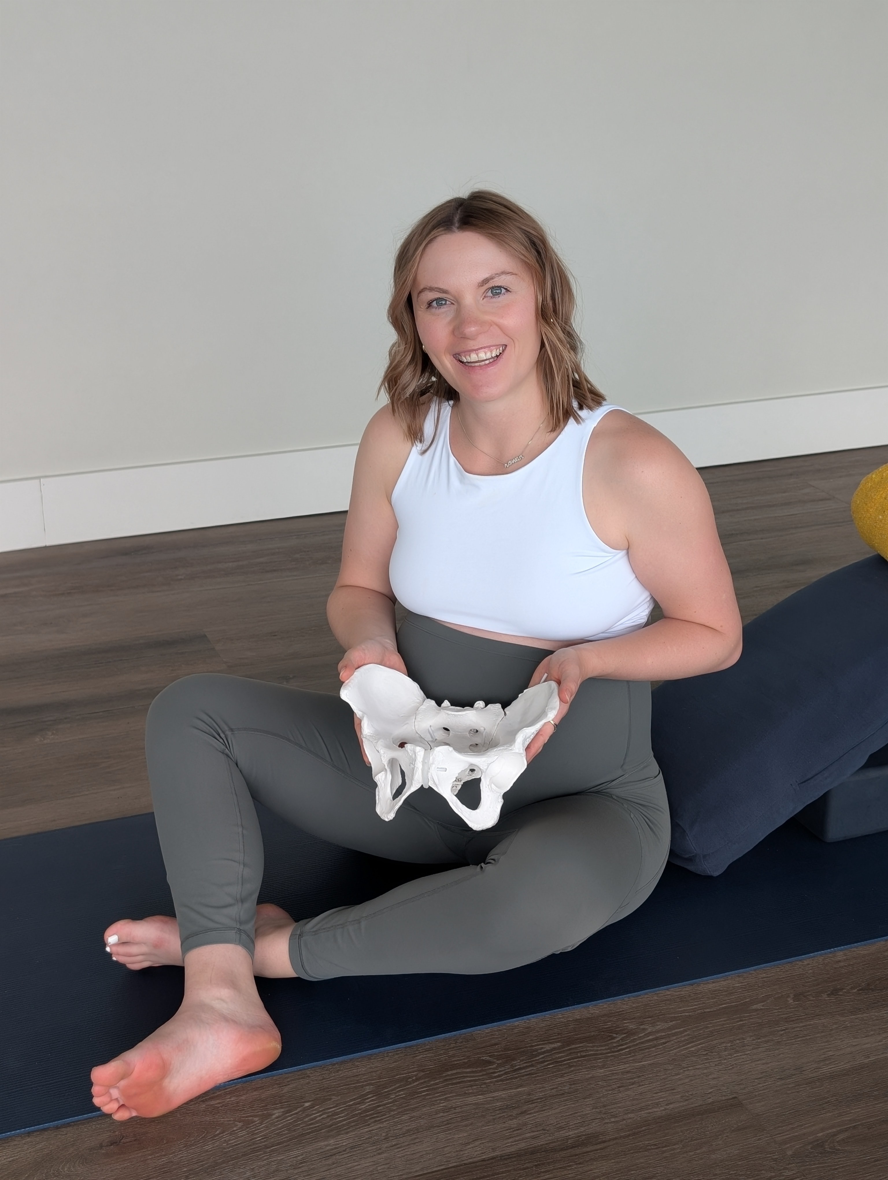 Woman sitting on yoga mat holding a pelvis model, smiling, in a room with wooden floor and plain wall.