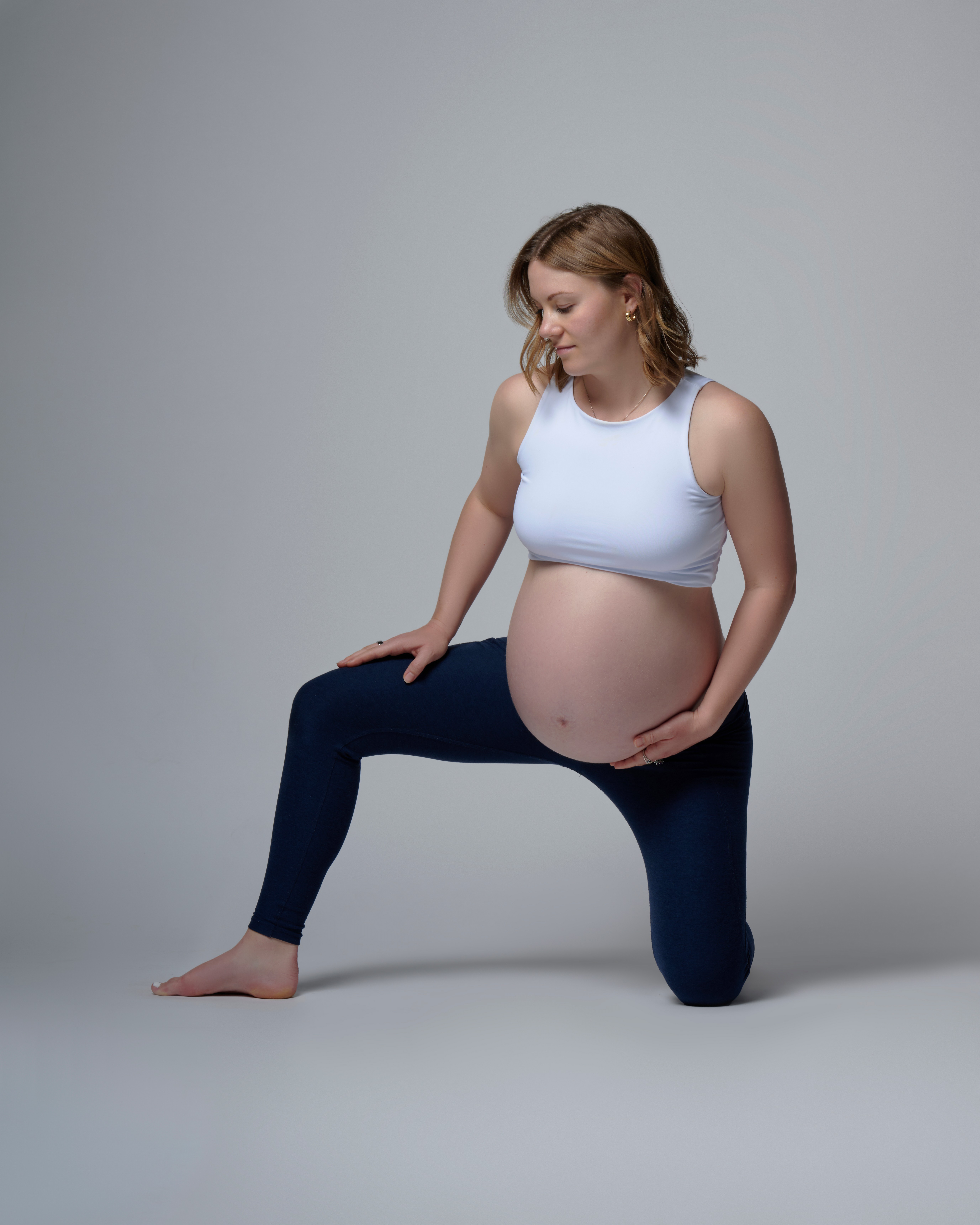 Pregnant woman kneeling with one knee on the ground, hand on her belly, wearing a white top and dark pants.