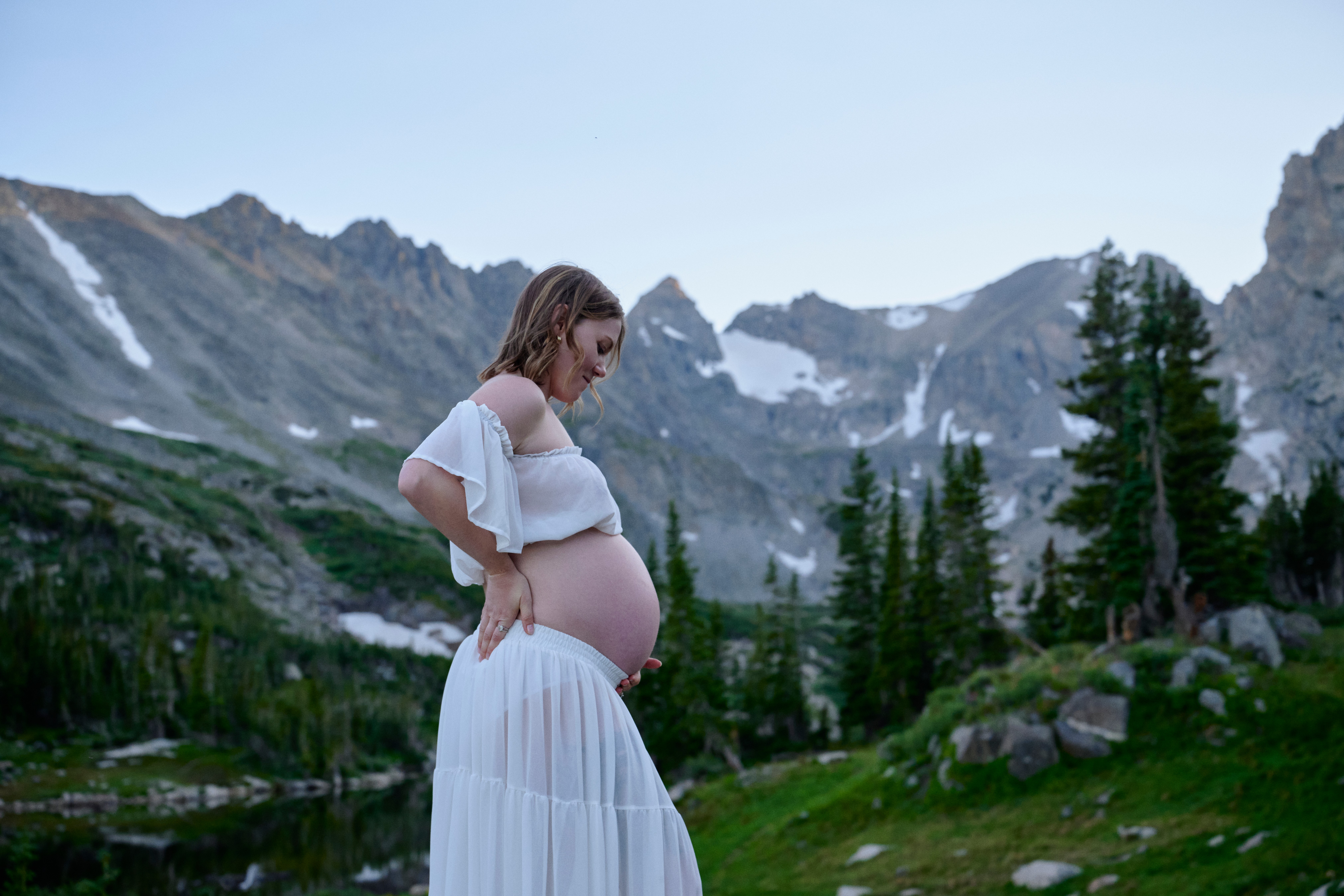 Pregnant woman standing outdoors in a mountainous landscape with trees and snow-capped peaks, looking down at her belly.